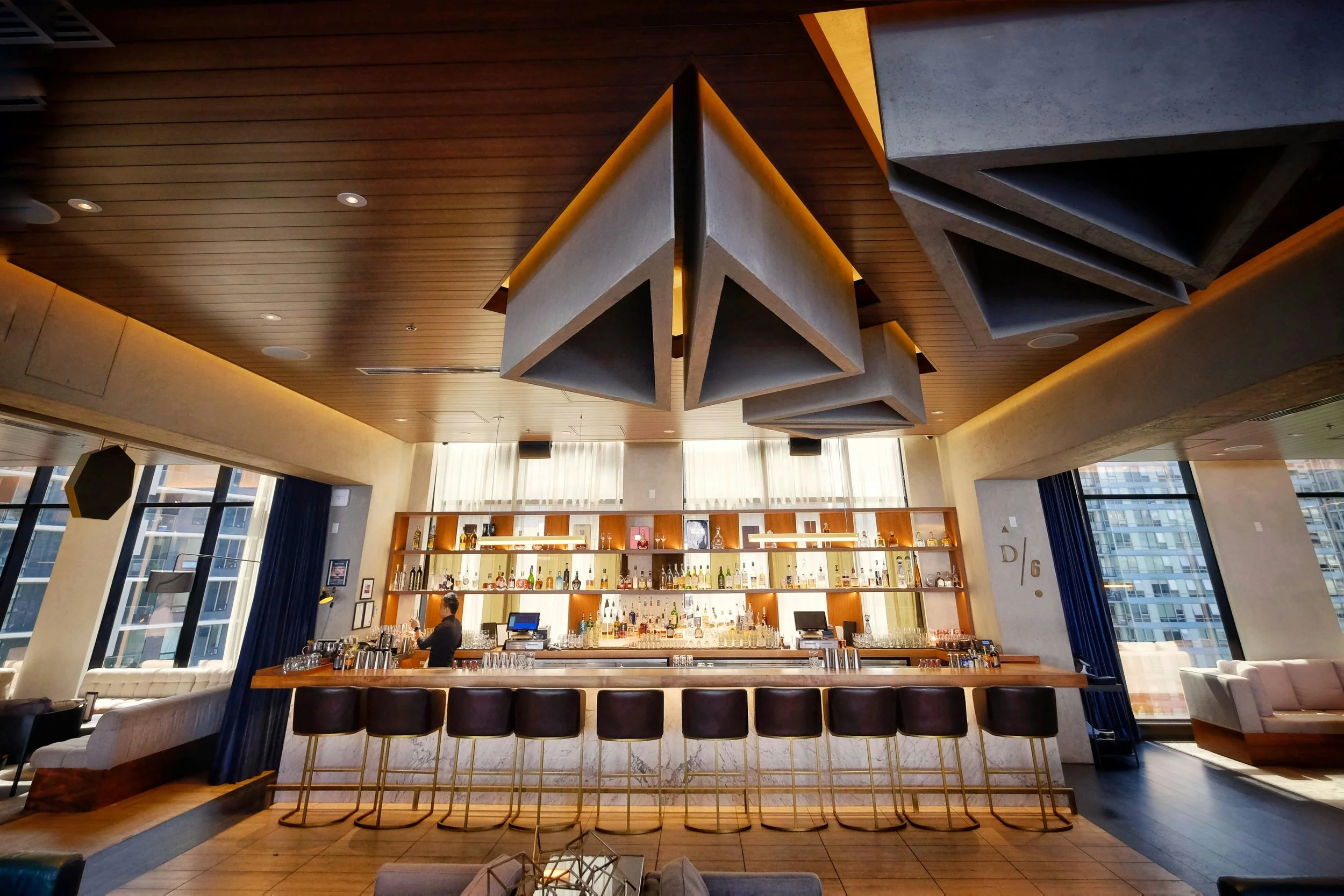 Modern bar interior with a high counter, bar stools, and a well-stocked back bar with liquor bottles, illuminated shelves, and a bartender preparing a drink. Large windows with blue curtains let in natural light.