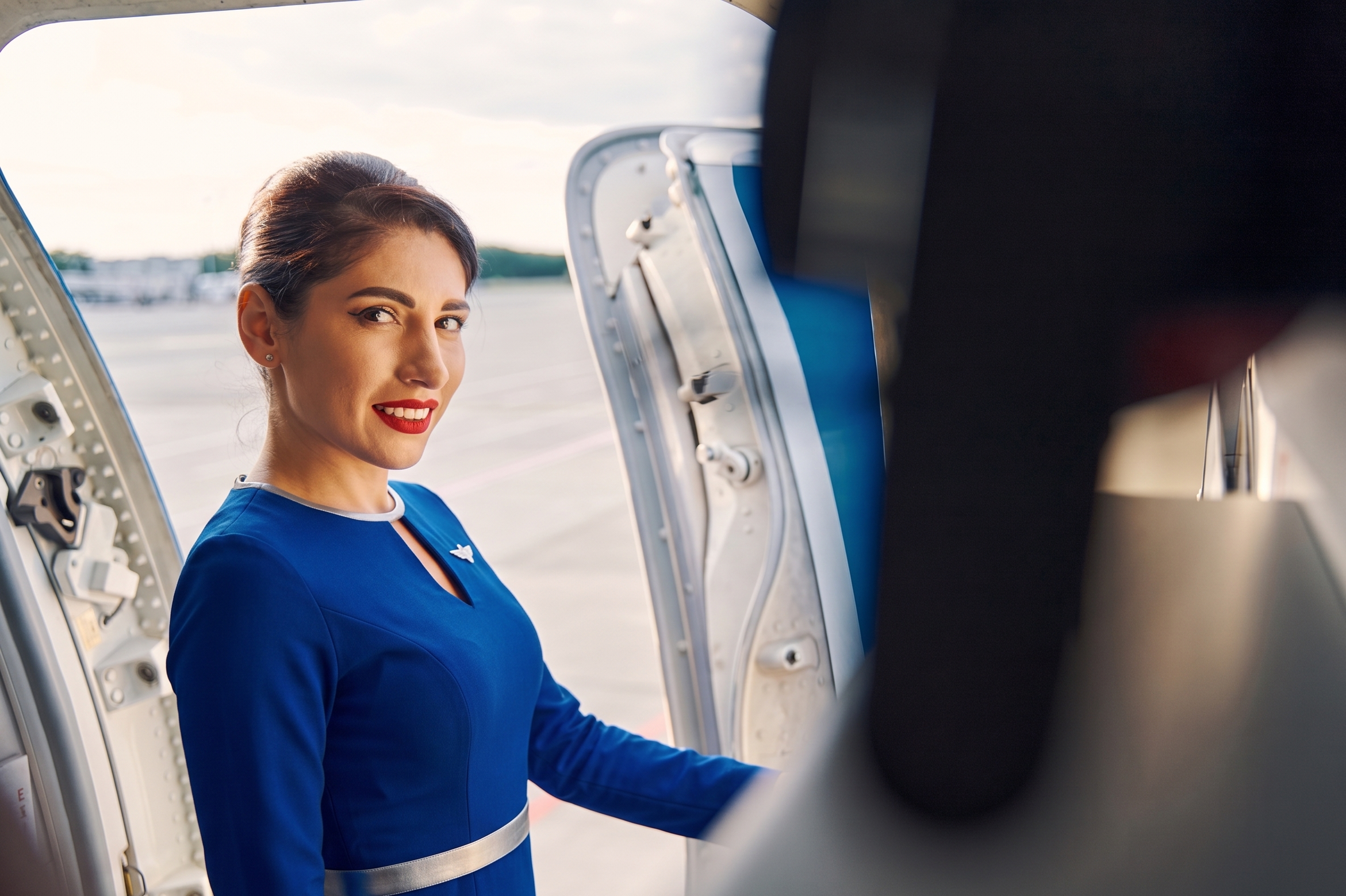 Una azafata con maquillaje y sonrisa, vestida con un uniforme azul, mira hacia la cámara desde la entrada de un avión.