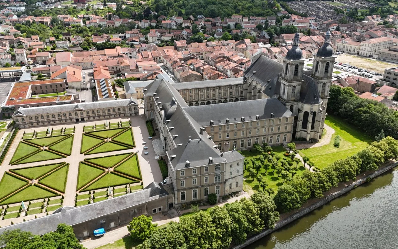 Abbaye des Prémontrés à Pont à mousson : Vue aérienne d'un grand bâtiment historique avec deux clochers, un jardin bien entretenu avec des motifs géométriques, et une rivière à proximité, dans une zone urbaine densément peuplée.