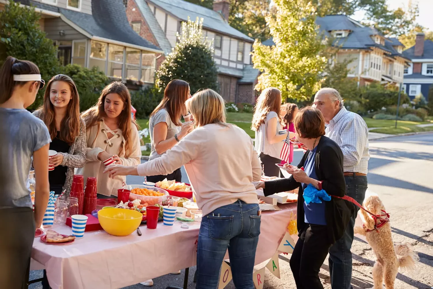 Une fête de quartier ou une réunion communautaire en plein air avec plusieurs personnes autour d'une table garnie de nourriture, dans un quartier résidentiel au coucher du soleil, avec des maisons et des arbres en arrière-plan.
