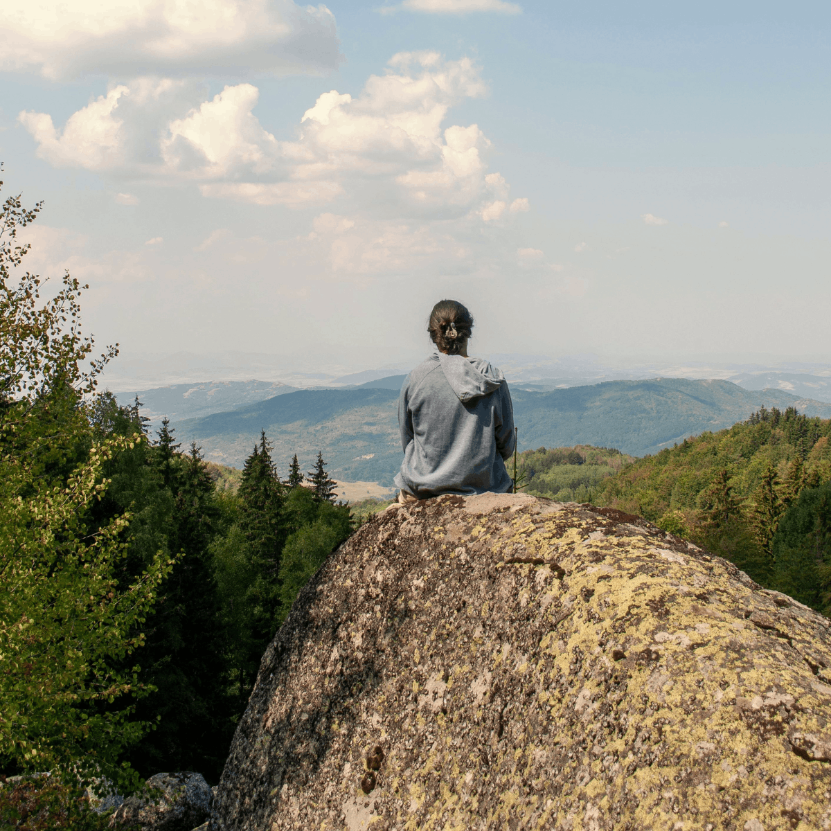 Person sitting on a large rock overlooking a valley with trees and mountains in the distance under a partly cloudy sky.