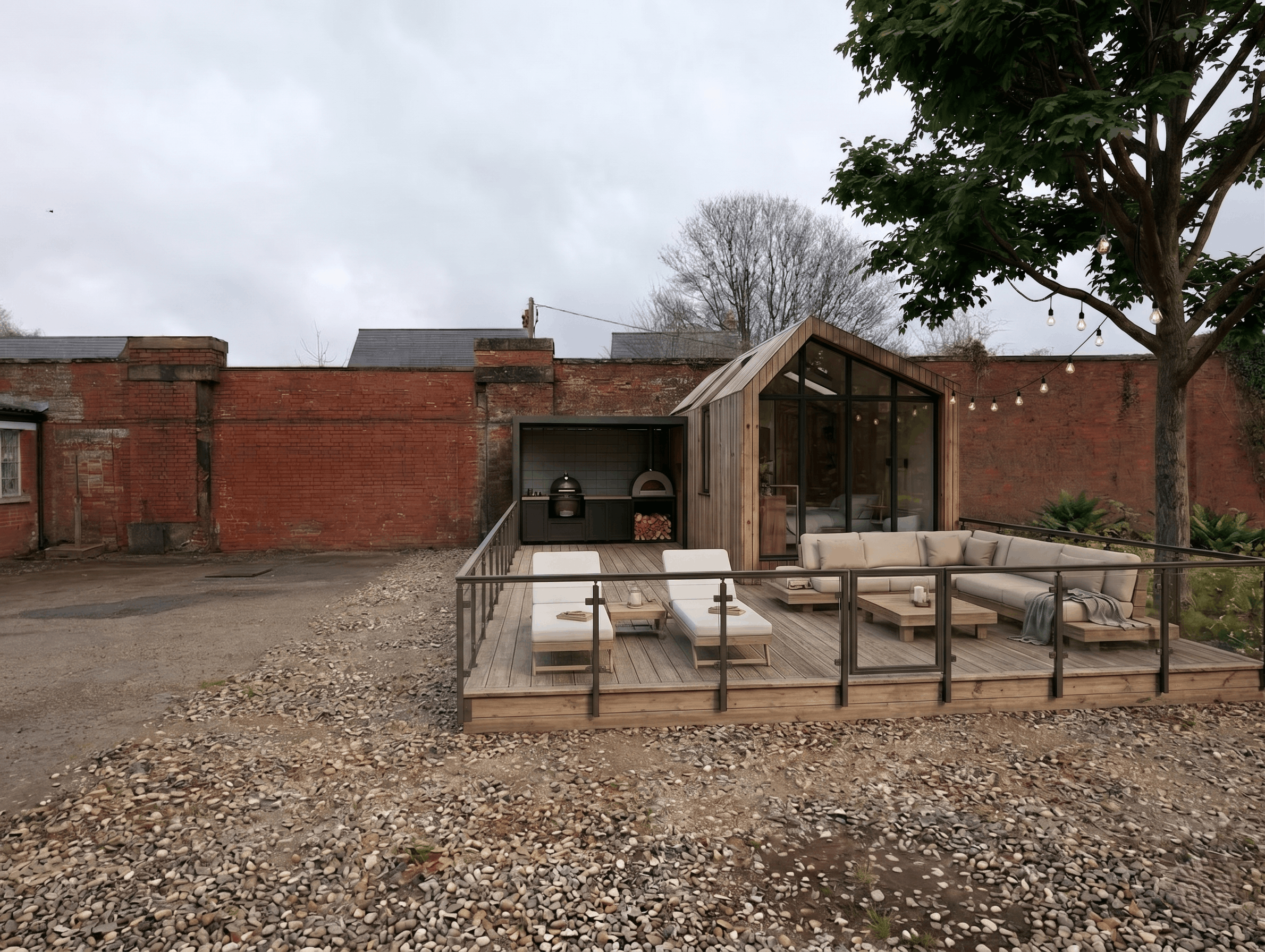 The SOMR Spaces showroom area with a wooden deck, outdoor sofa, coffee tables, string lights, a large tree with green leaves, and a red brick wall in the background on a cloudy day.