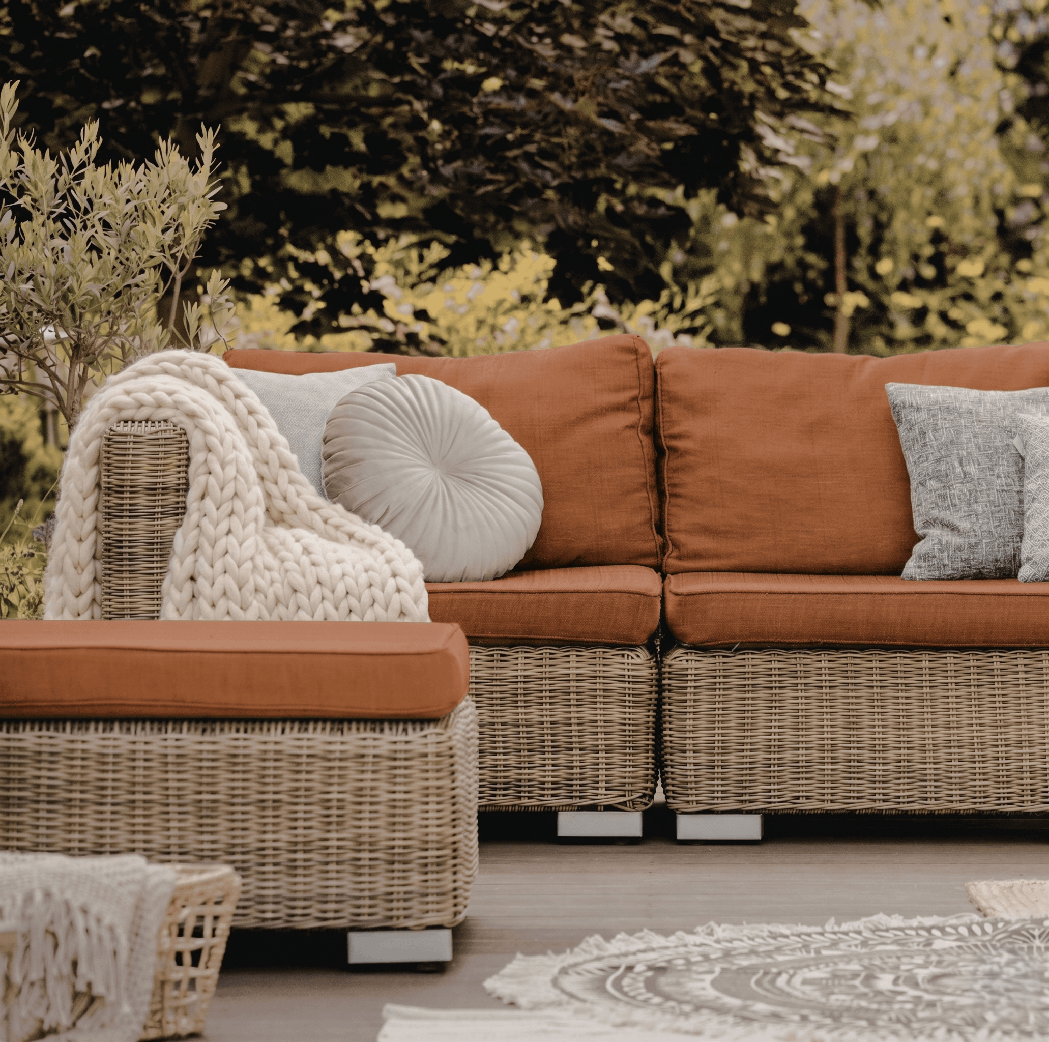 A cozy outdoor seating area with a red sofa, decorative pillows, a knitted blanket, wicker side table, and a dining table with a round rug beneath, set against a background of trees and greenery.