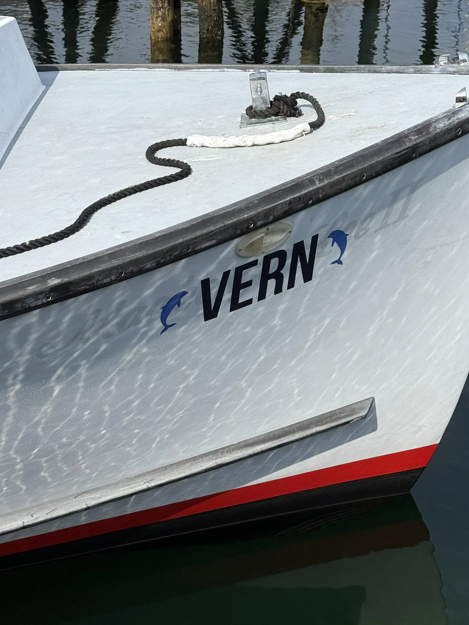 Close-up of the bow of a boat docked in water, showing a black rope and a white cloth tied to a metal cleat on the boat's surface, with water and wooden posts in the background.