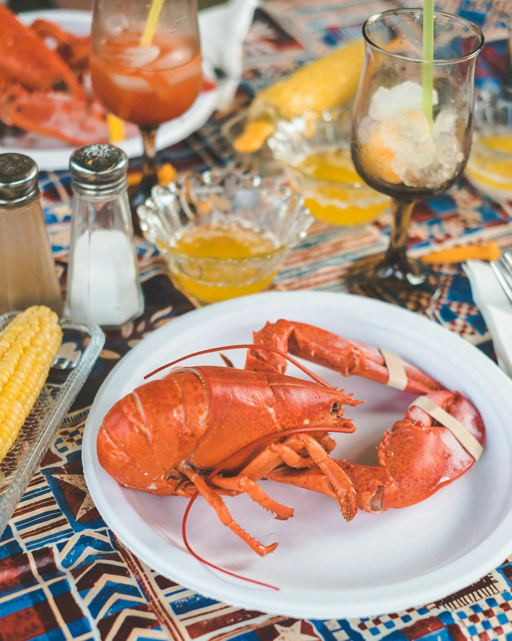 Lobster on a white plate with corn, lemon, and seafood with a colorful tablecloth.