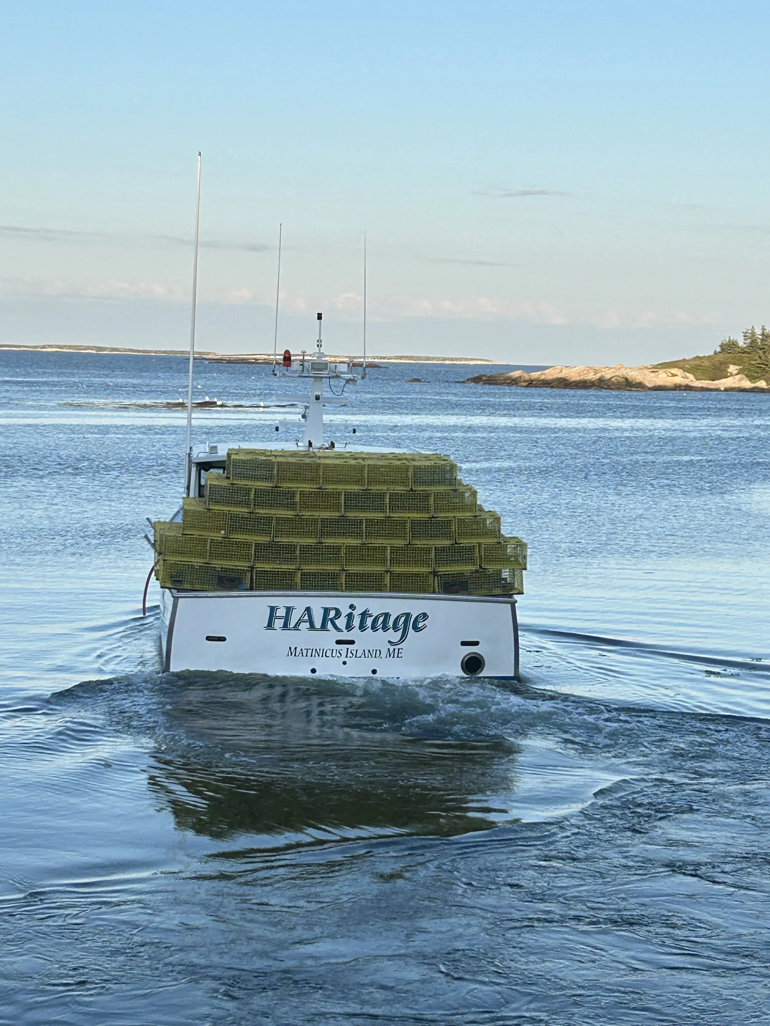 A boat on the water with a sign reading 'HARitage' and 'MATINICUS ISLAND, ME' attached to it, with land and sky in the background.