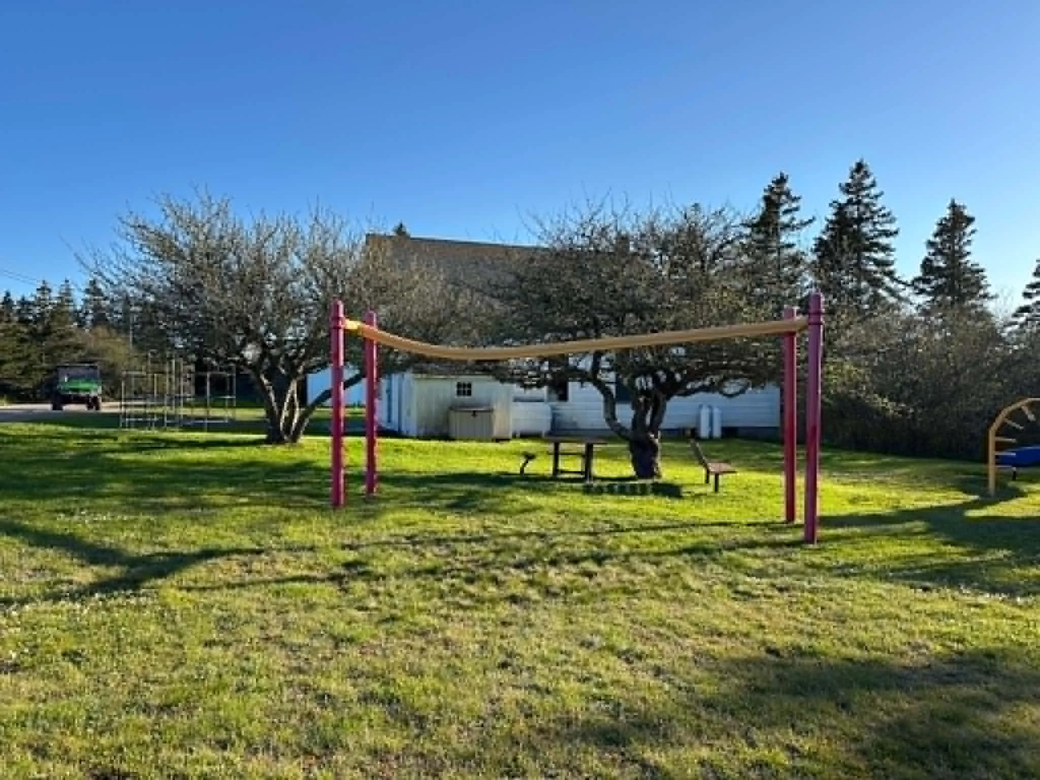 A playground with a swing set, benches, trees, and a white building in the background, under a bright blue sky.