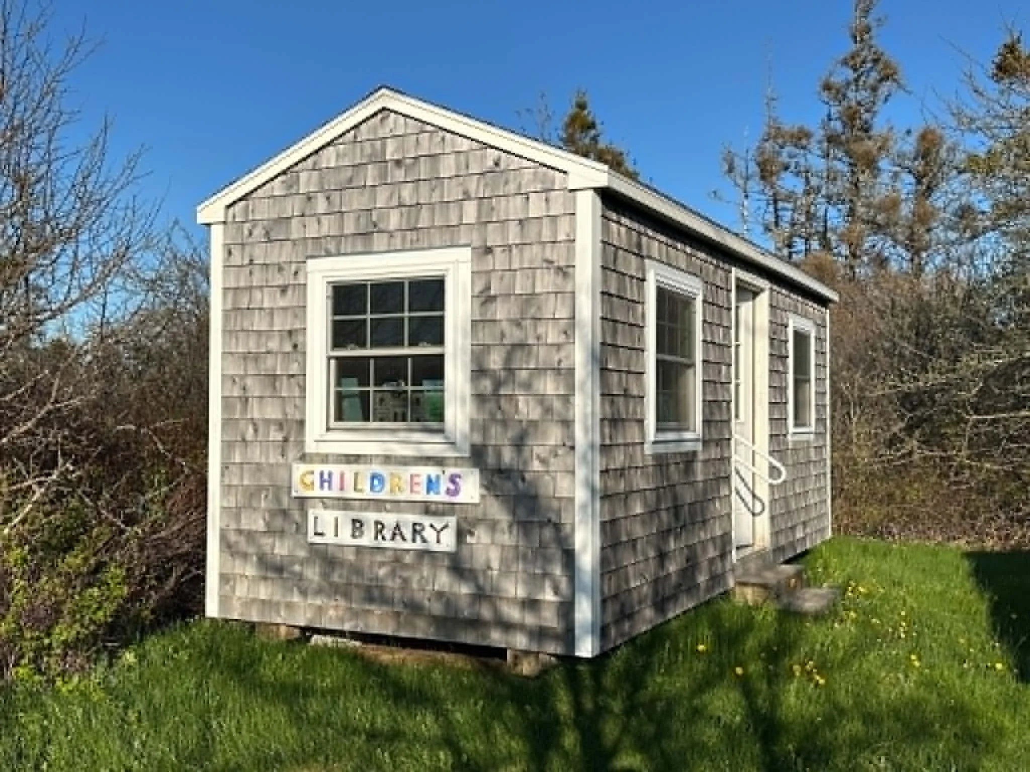 Small wooden building with shingle siding and white trim, labeled as a children's library, surrounded by grass and trees, under a blue sky.