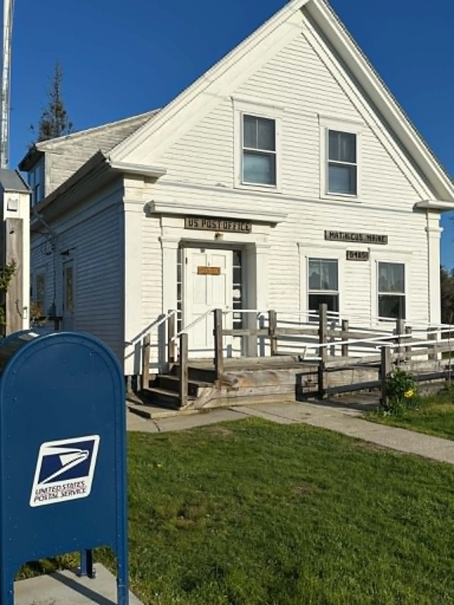 White wooden house with a gable roof, two first-floor and two second-floor windows, a front door with stairs and a ramp, and a blue mailbox in the foreground on a sunny day with a clear blue sky.