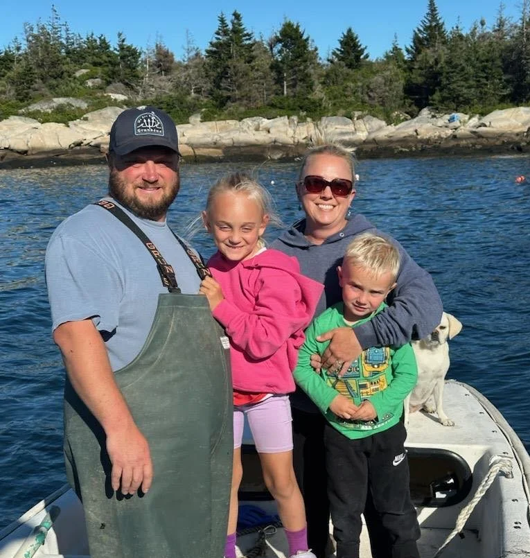 A family of four, with a dog, standing on a boat near the water, smiling at the camera, with trees and rocks in the background.