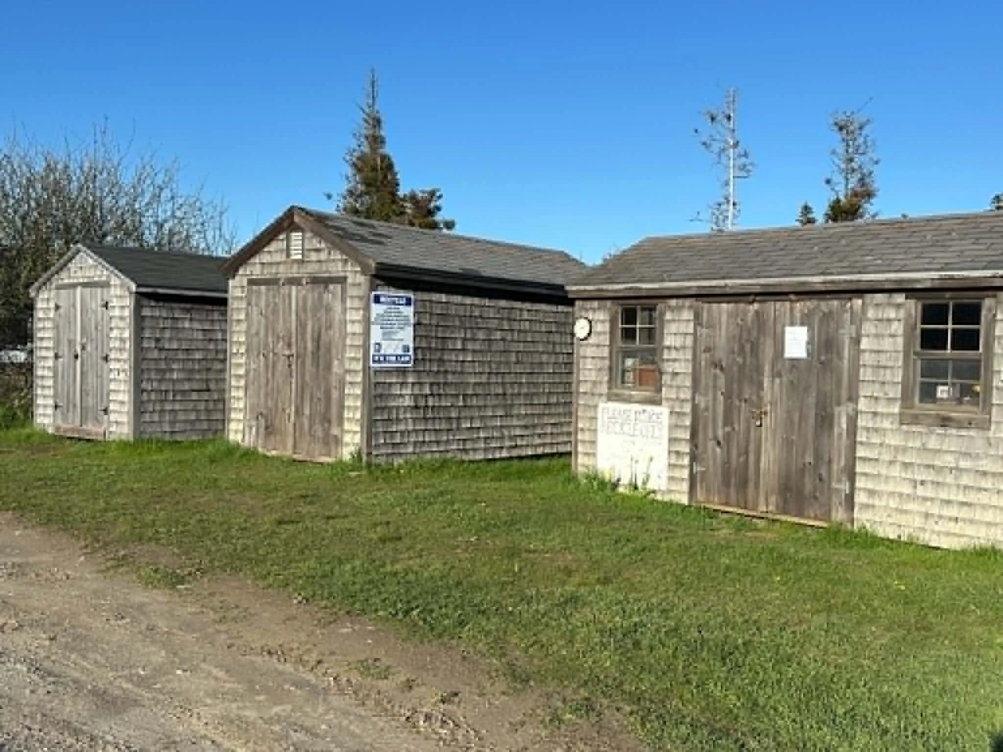 A small wooden building with a shingled roof and windows, surrounded by a grassy area and a gravel path, under a clear blue sky with some leafless trees in the background.
