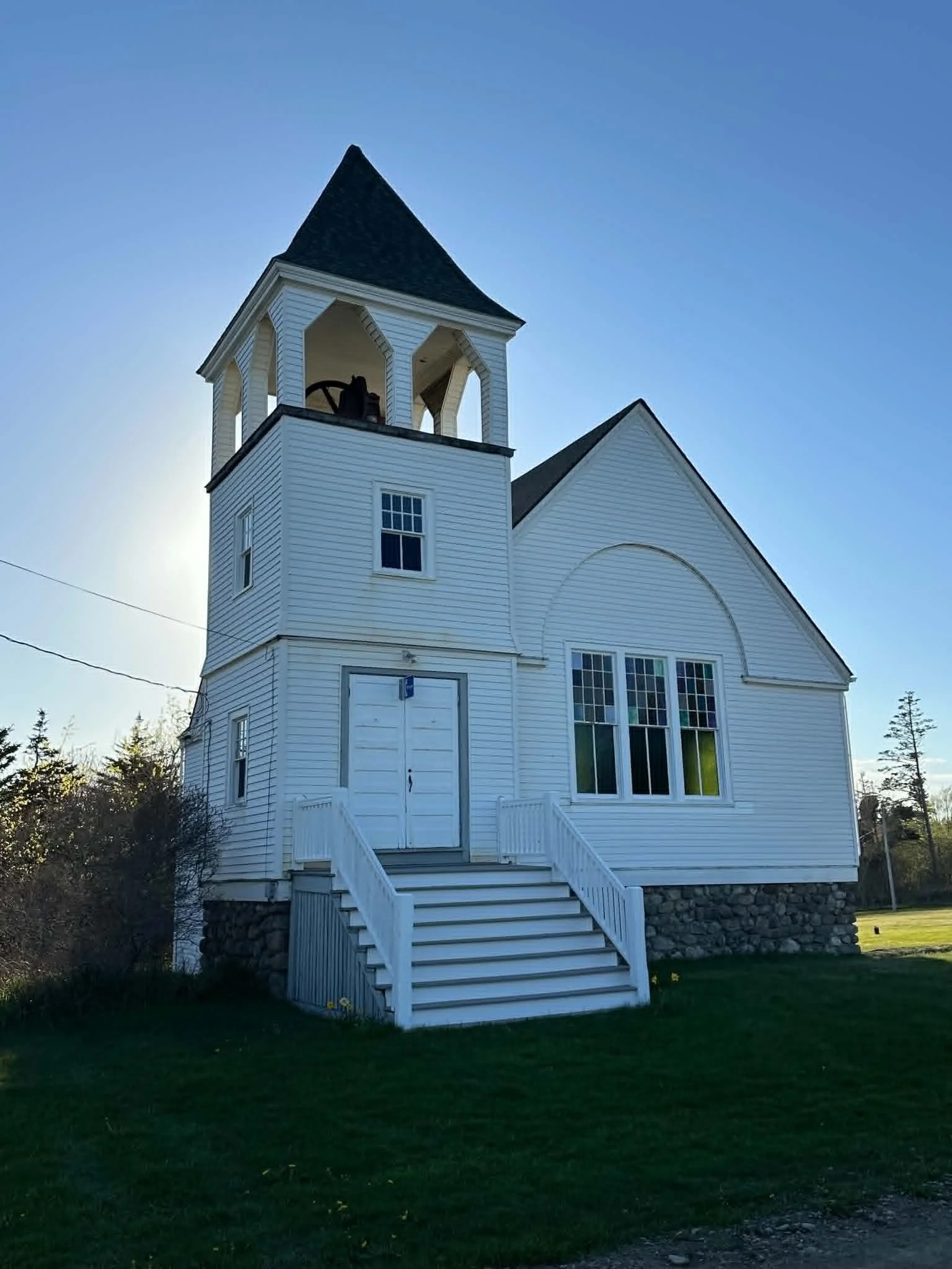 A white wooden church with a steeple, stairs leading up to the entrance, large windows, and a stone foundation under a clear blue sky.