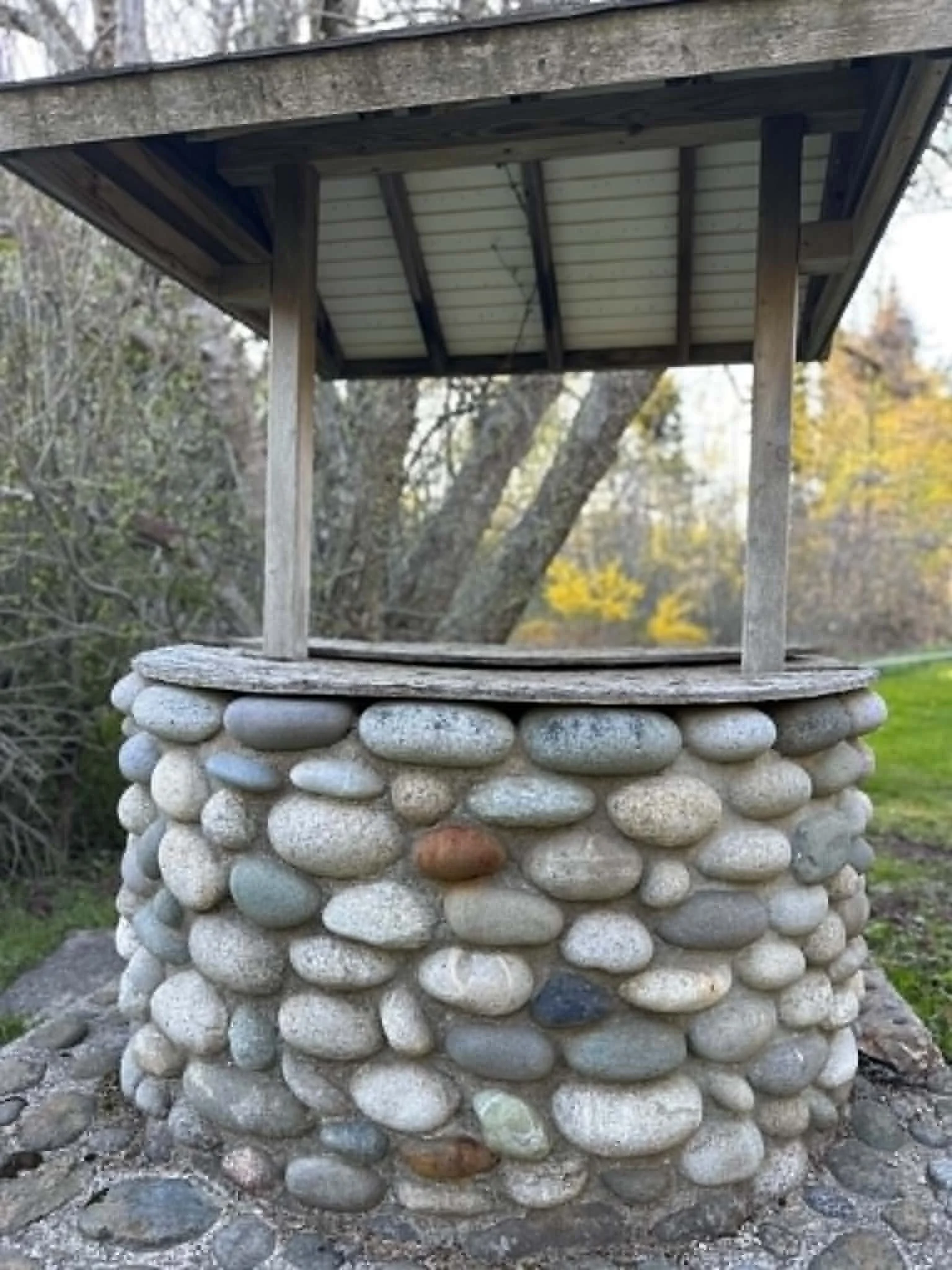 A small stone well with a wooden roof and stone pillar base, surrounded by grass and trees in an outdoor setting.