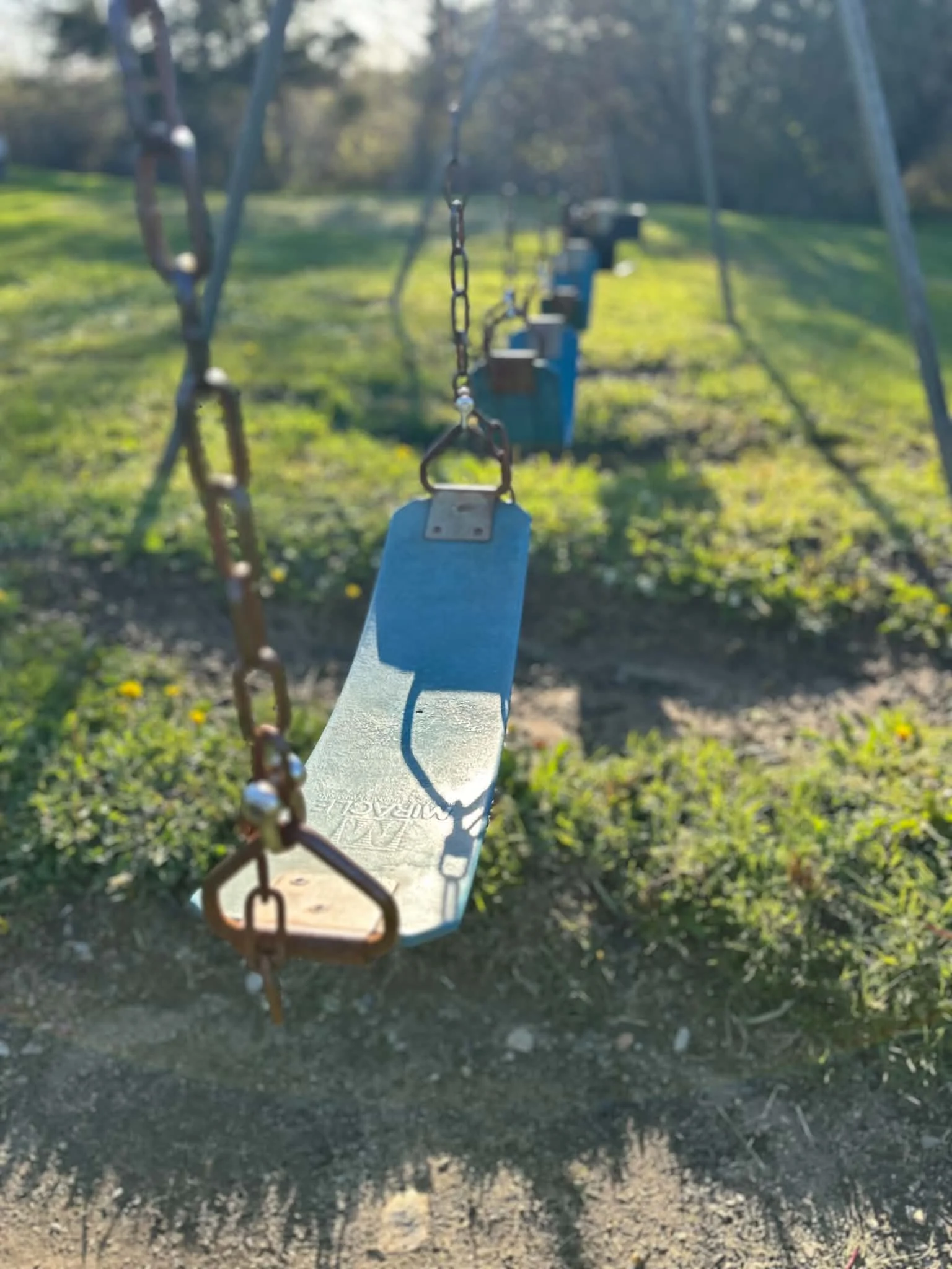 Close-up of a swing chain and seat at a children's playground, with green grass and trees blurred in the background.