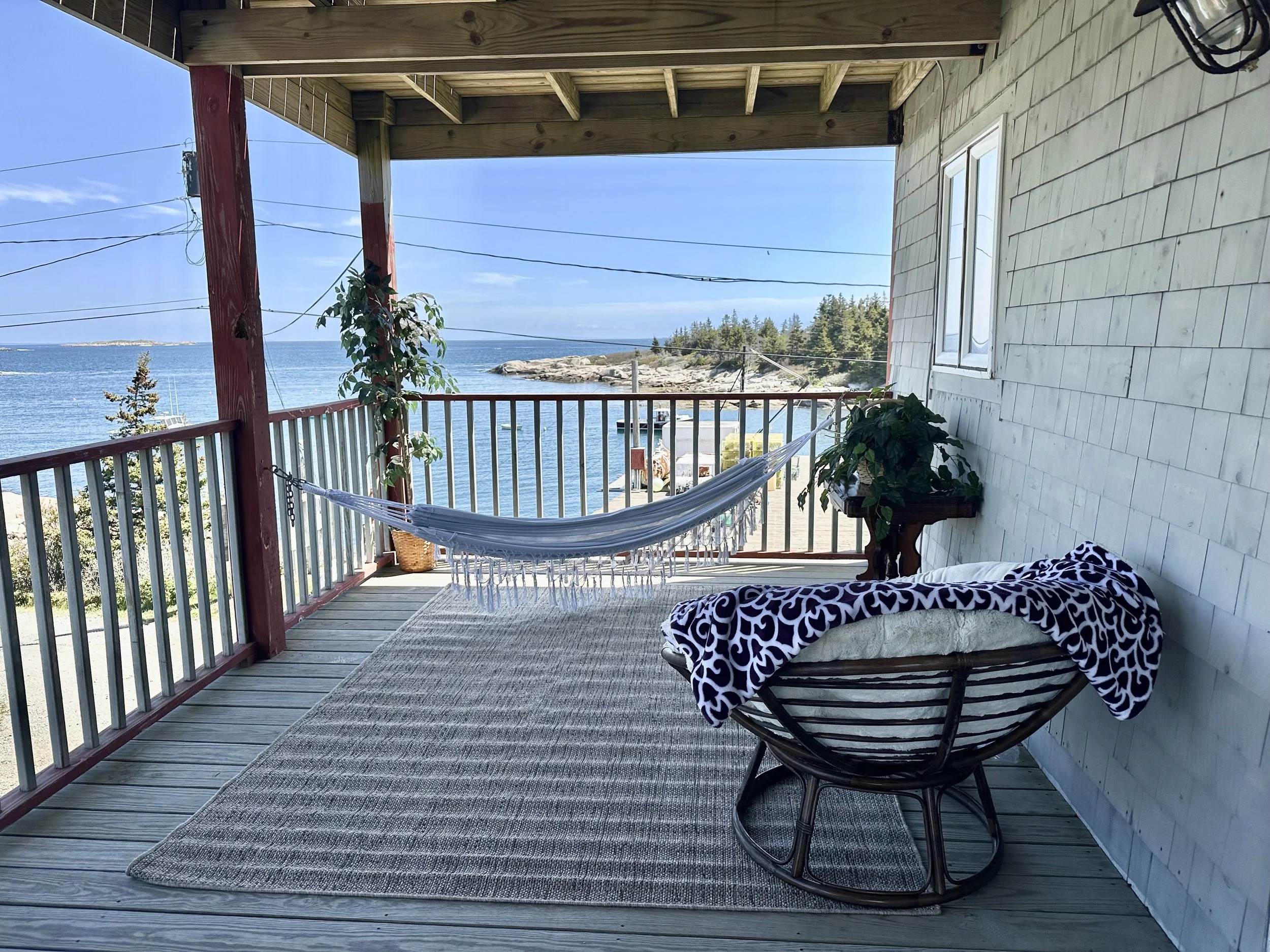 A balcony overlooking a body of water with trees in the background. The balcony has a hammock, a plant, and a small table with a patterned cloth, framed by a wooden railing and ceiling.