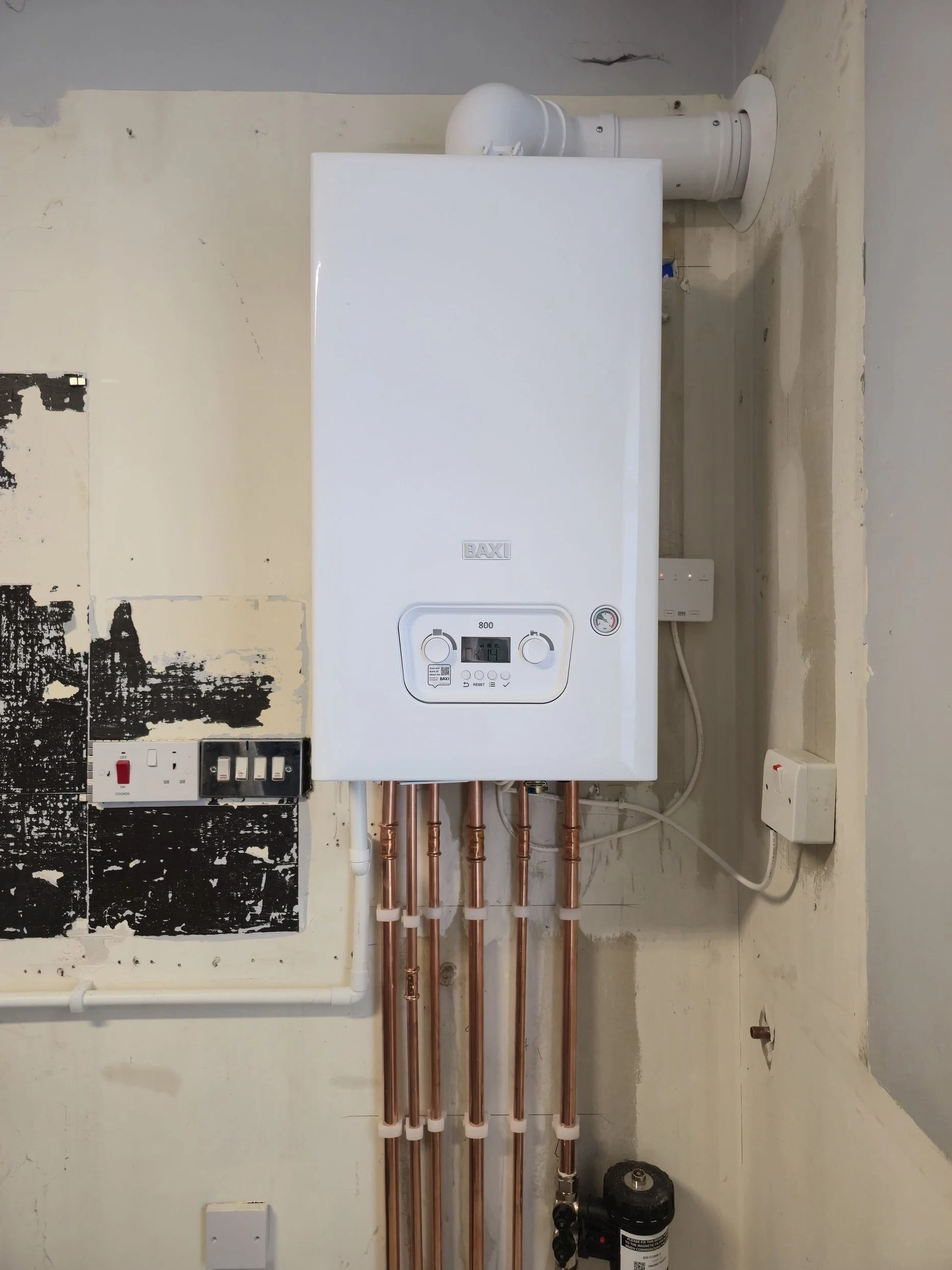 A white boiler with digital display and knobs, copper pipes connected below, mounted on a wall in a utility room or basement.