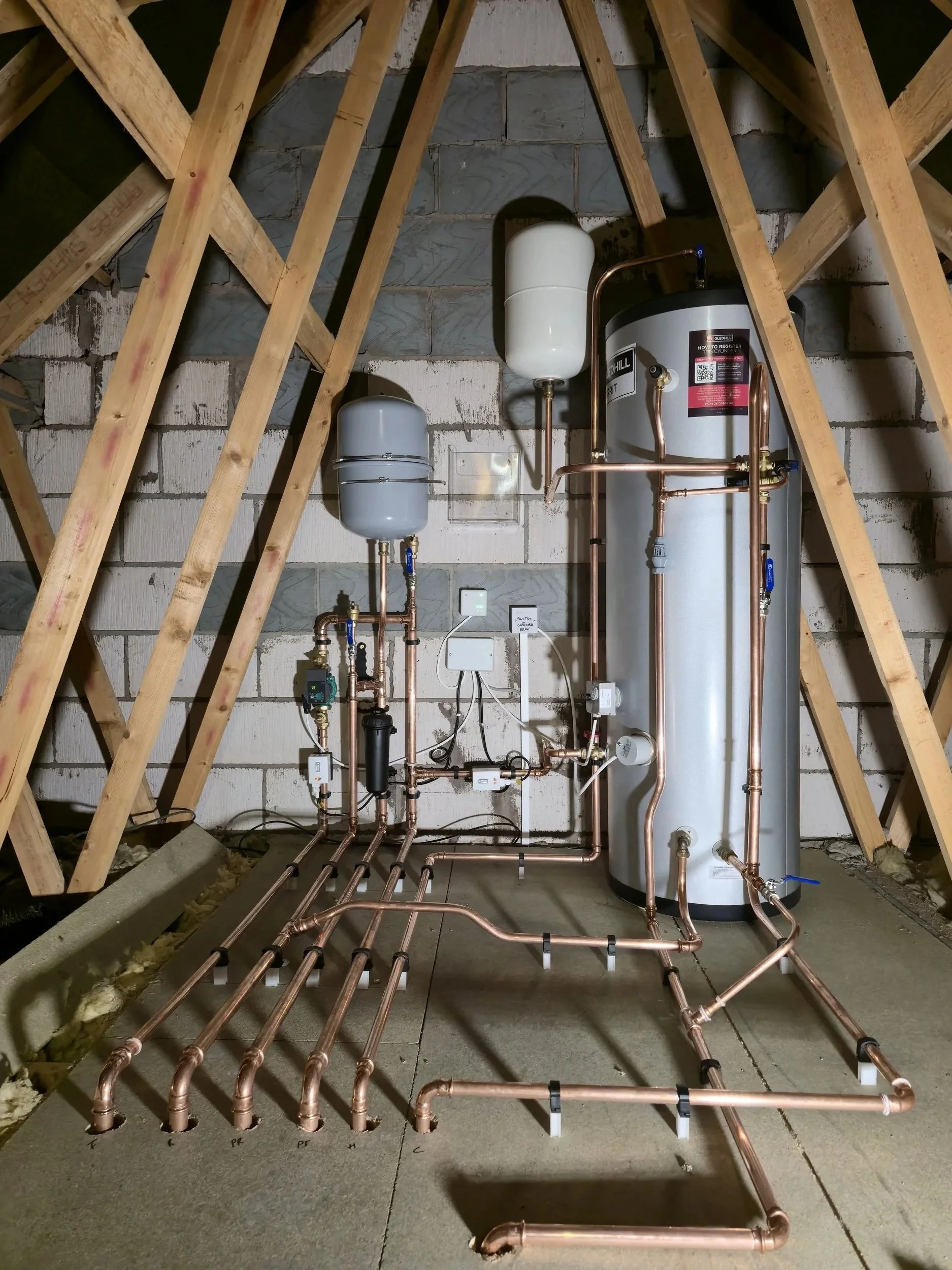 A mechanical room with plumbing pipes, a water heater, and an expansion tank installed on a brick wall, with wooden framing above.