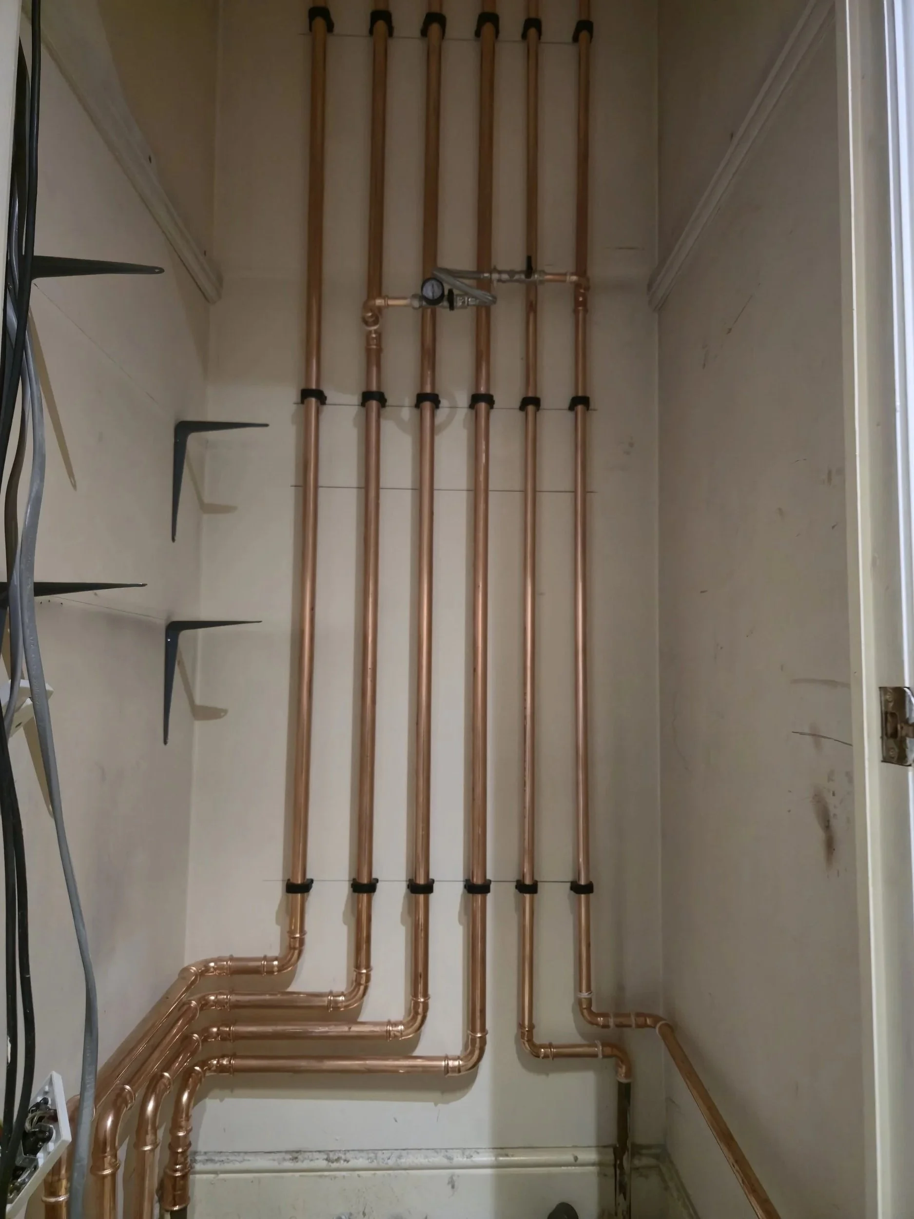 Copper plumbing pipes with a pressure gauge and fittings inside a small utility closet or cabinet.