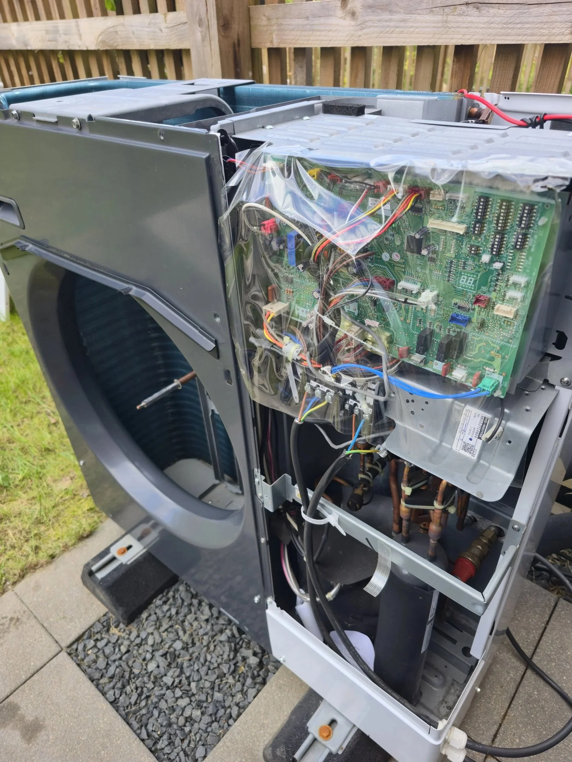 Open electrical control box with circuit boards, wiring, and components inside, on a paved outdoor surface.