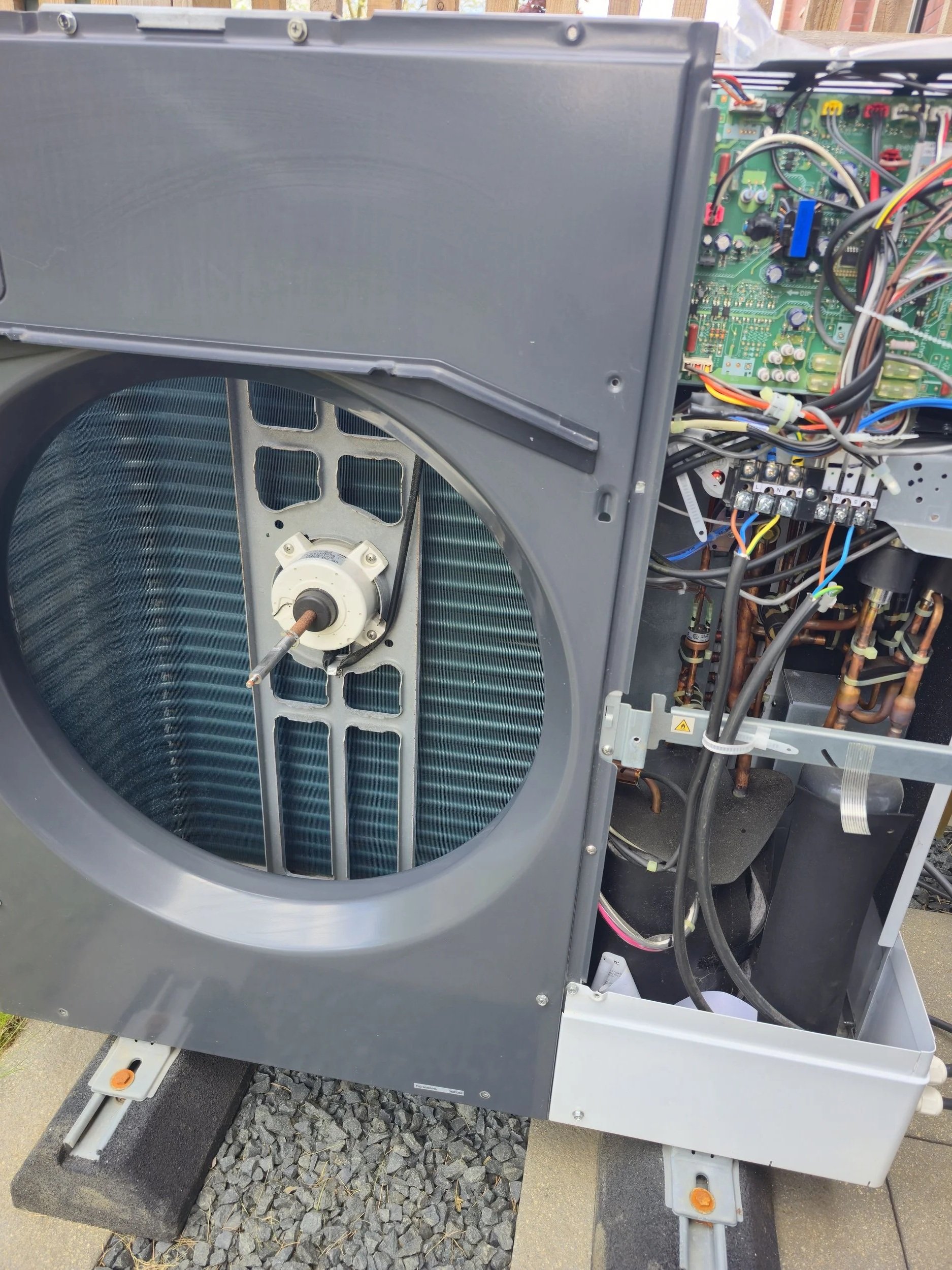Open washing machine with exposed electronic components and wiring, sitting outdoors on concrete and gravel.