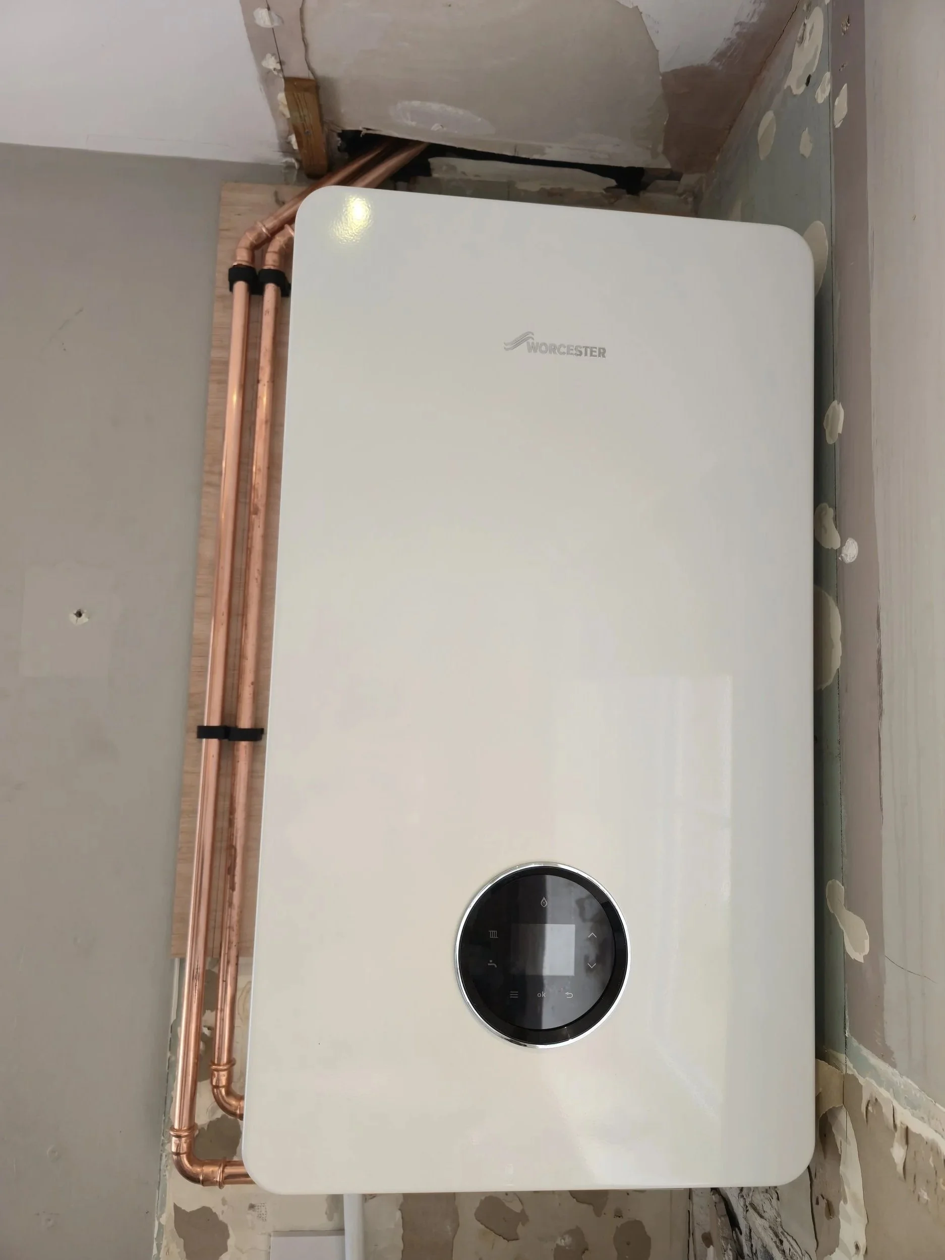 A white Worcester boiler mounted on a wall with exposed copper pipes nearby, in a room with unfinished drywall and wall patches.