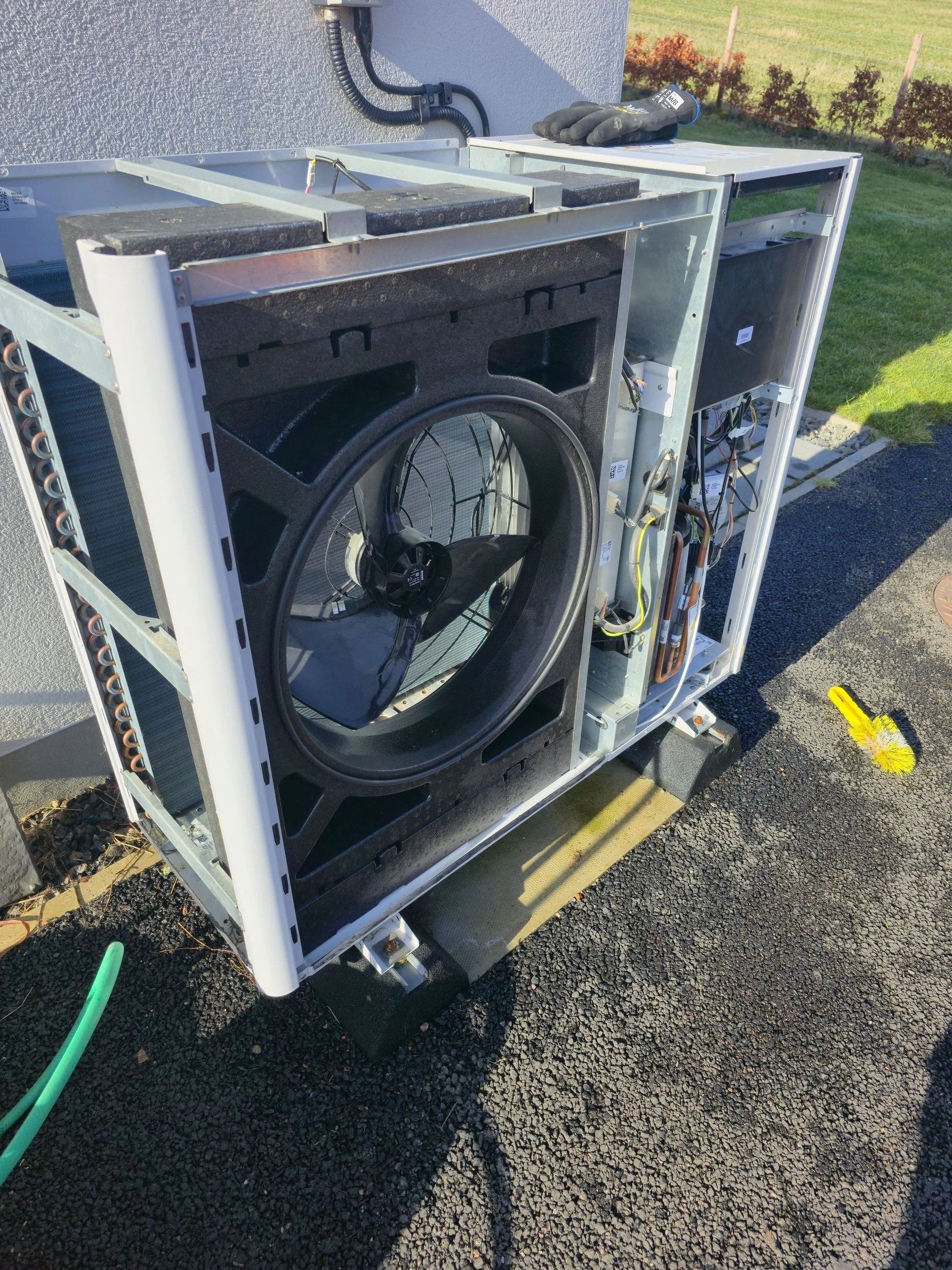 The image shows an outdoor unit of an air conditioning system with the front panel removed, exposing the internal components like the fan, coils, and wiring. A pair of work gloves and a brush are placed on top of the unit.