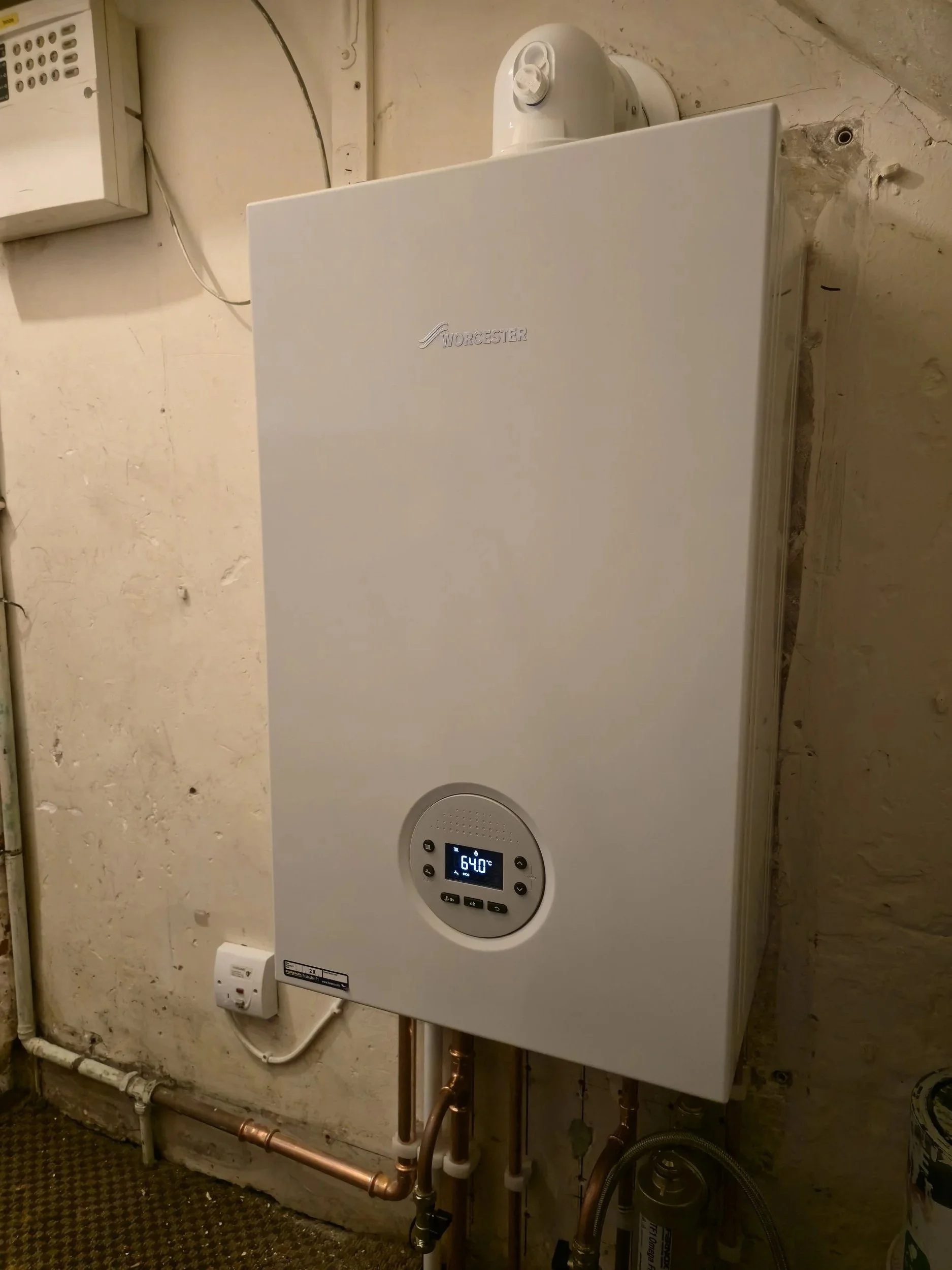 A white Worcester boiler mounted on a wall with pipes underneath, a digital display showing 64.0°C, and an electrical outlet with a plugged-in device.