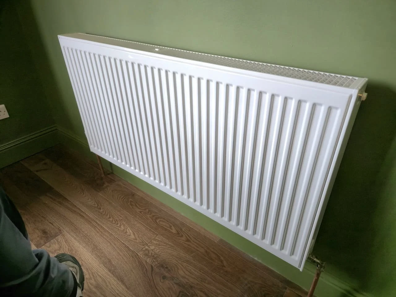 A white panel radiator mounted on a green wall, above a wooden floor.