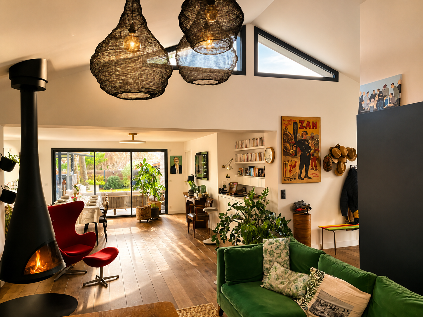 Maison individuelle – Réalisation – Cheminée centrale suspendue, fauteuil rouge et canapé en velours vert : dans ce séjour sous charpente, les verrières zénithales et les grandes baies coulissantes inondent de lumière un intérieur éclectique, chaleur