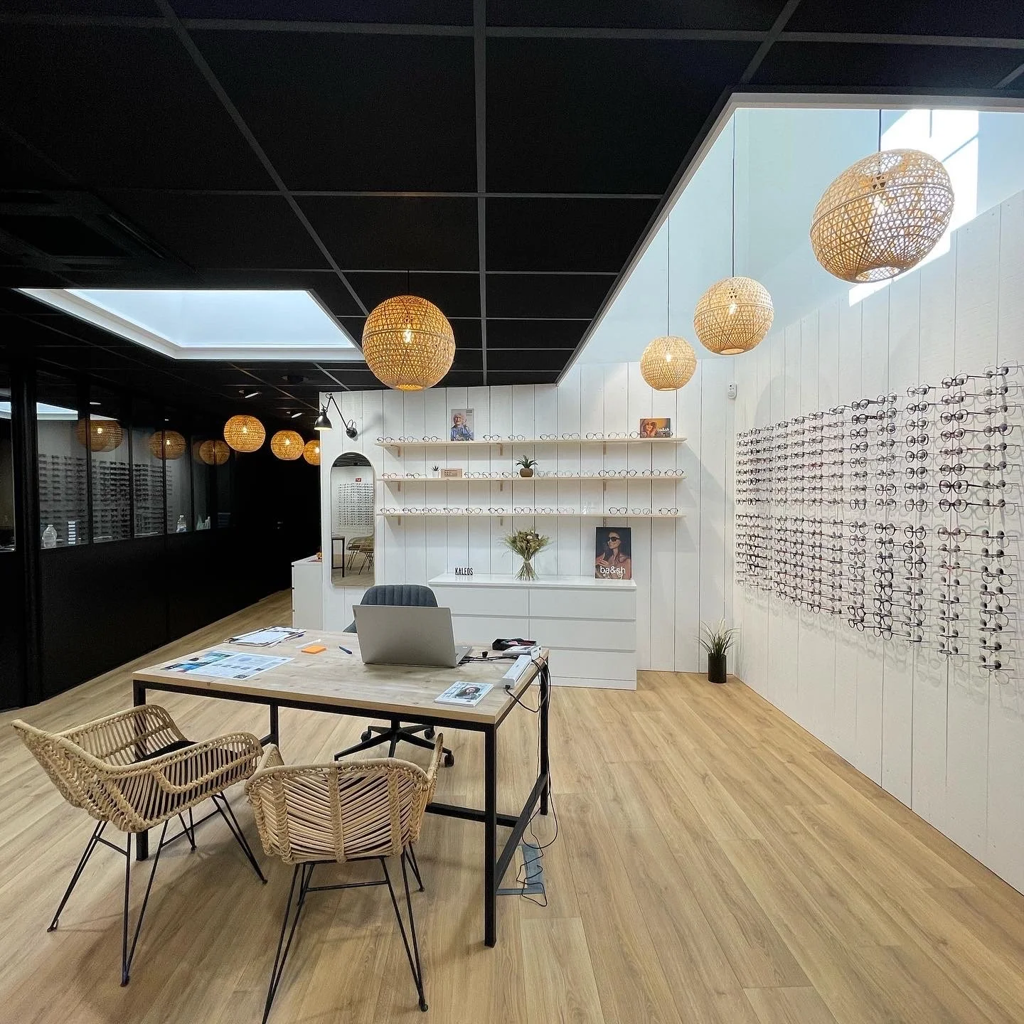 Plafond noir, suspensions en rotin doré et présentoirs muraux blancs : un contraste soigné qui met en lumière les montures dans une atmosphère chic et intimiste.