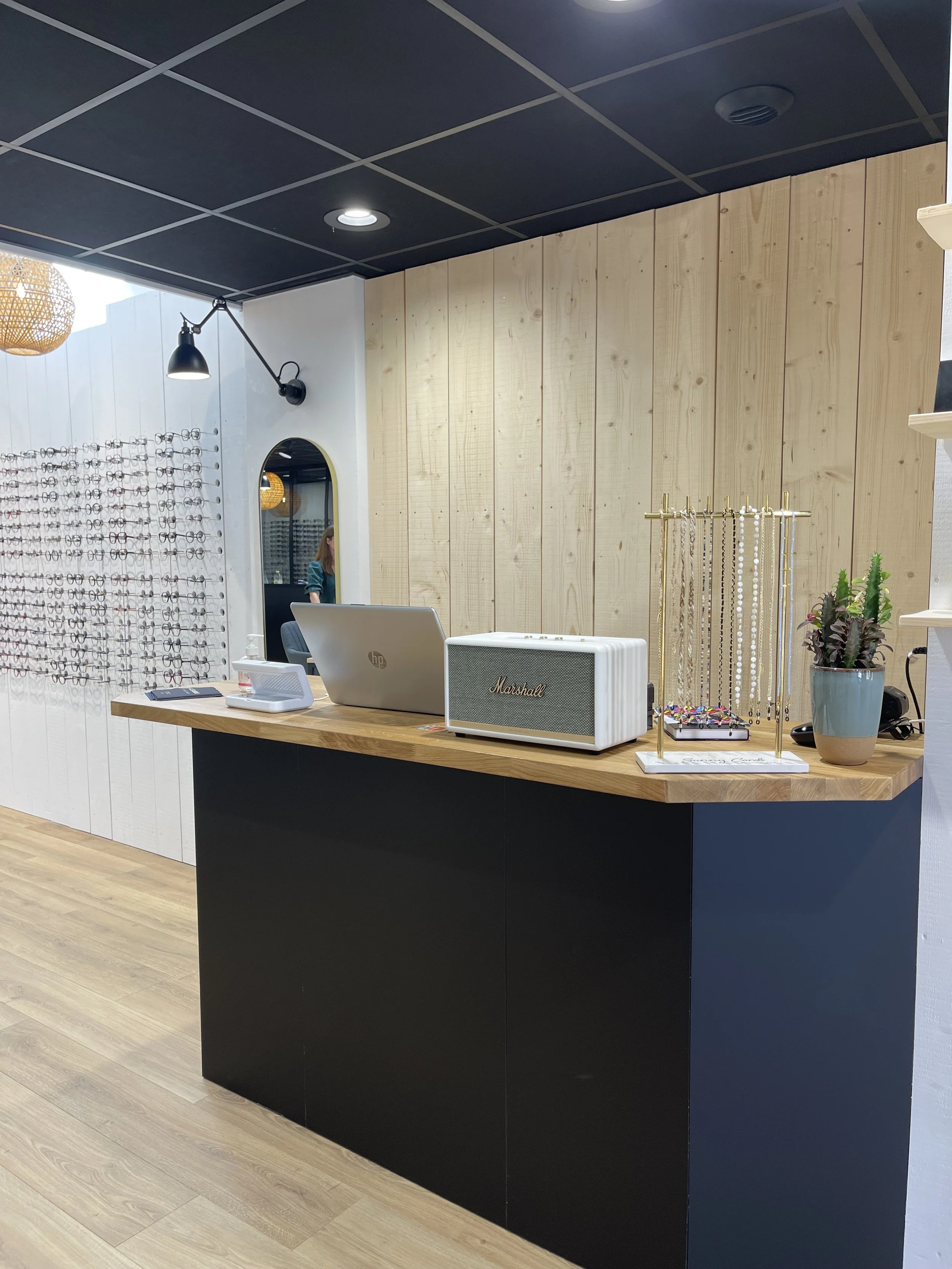 Boutique Optique – Réalisation – Comptoir bimatière en bois et noir mat, mur de montures et niche arquée : un espace d'accueil élégant qui allie fonctionnalité et identité visuelle affirmée.