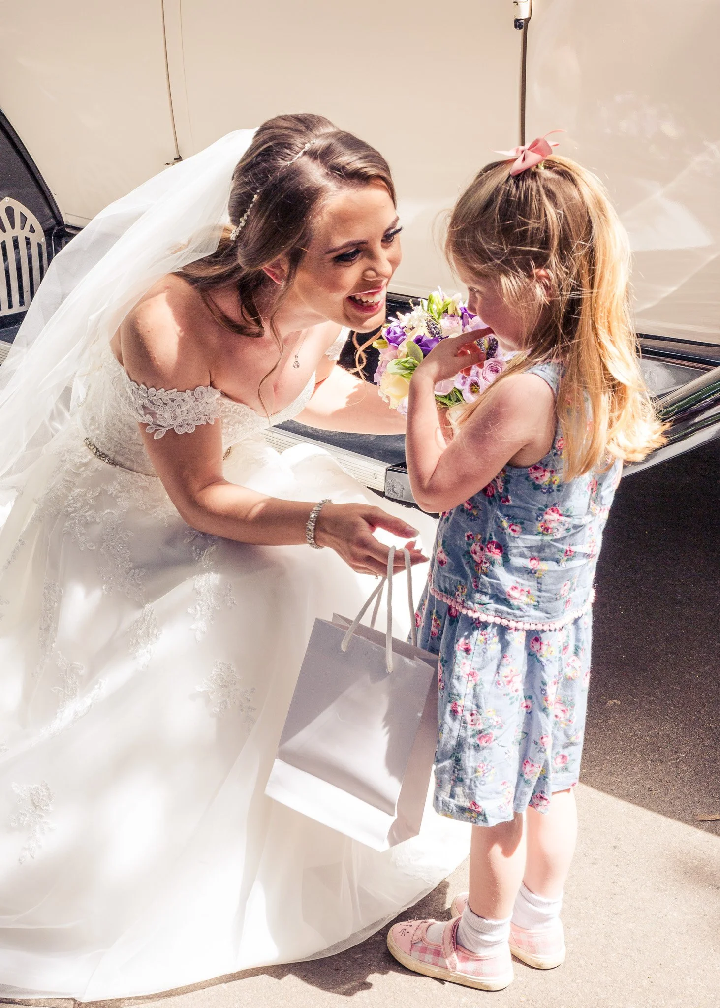 A young girl with blonde hair wearing a colorful dress, pink bow headband, and pink shoes stands next to a woman in a wedding dress holding a bouquet of flowers. The woman appears to be a bride, smiling at the girl as they interact outdoors on a sunn