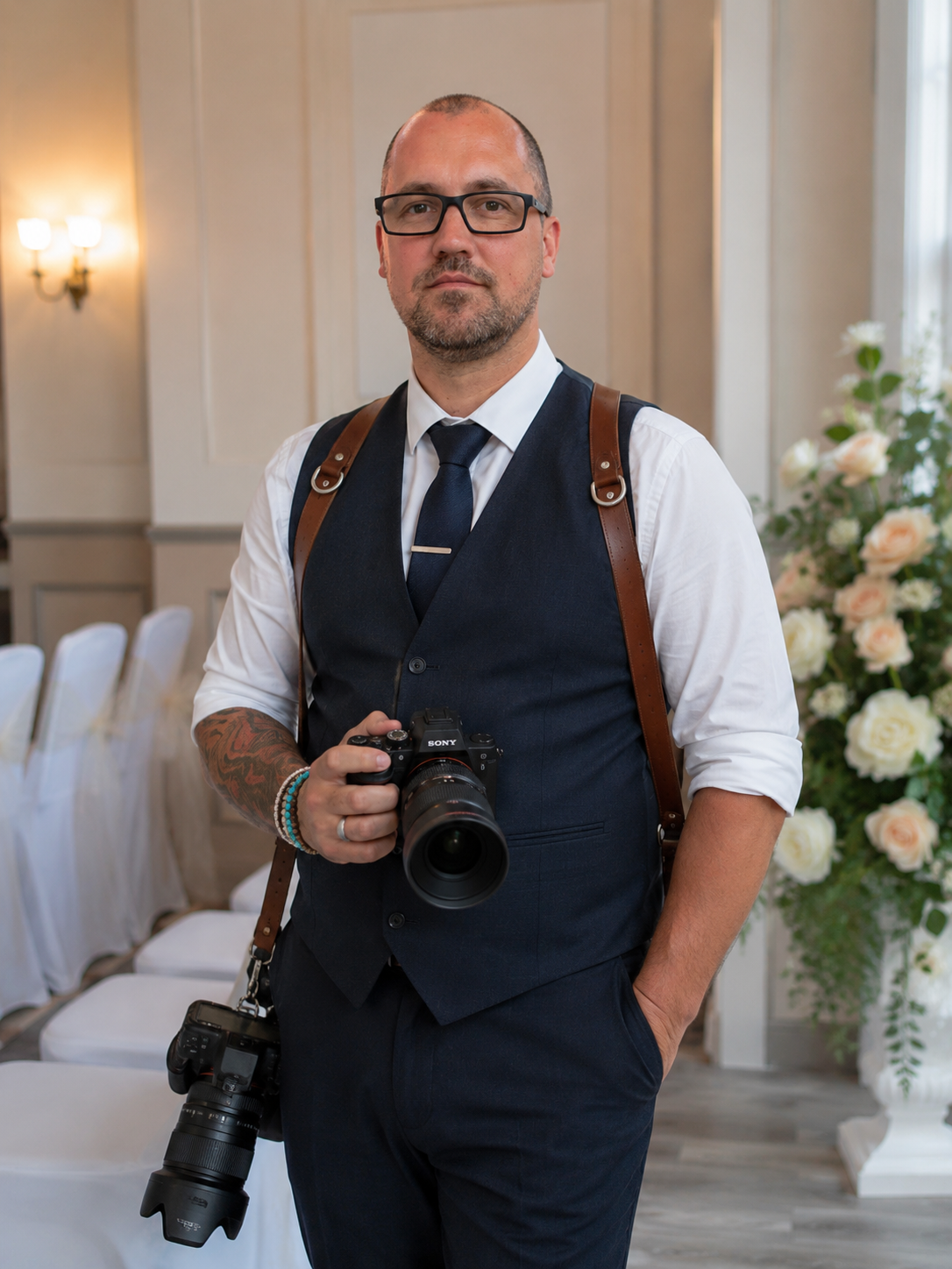 A man wearing glasses, a white shirt, and a navy vest holding a camera, standing in a decorated indoor space with floral arrangements.