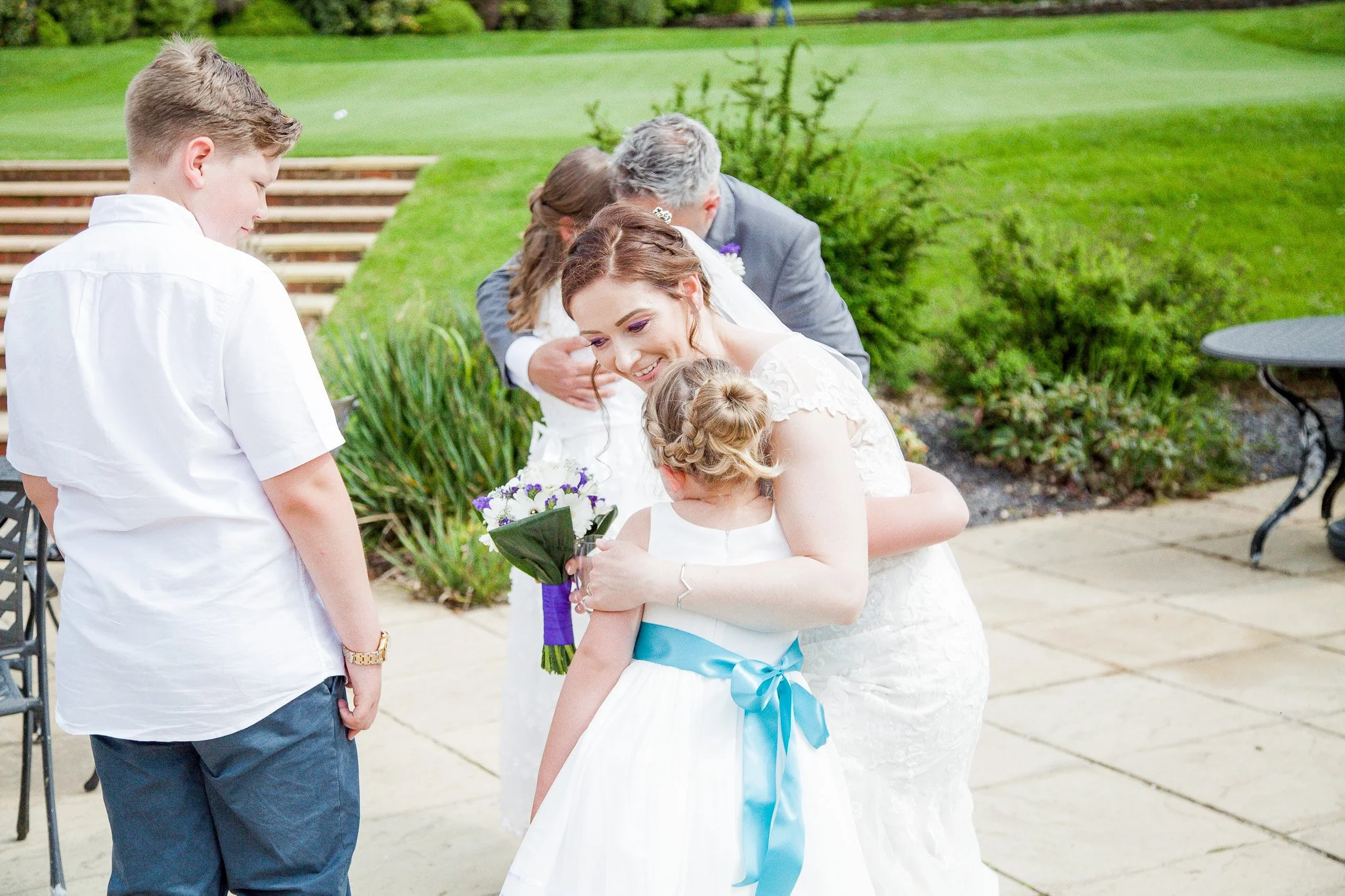 A woman in a wedding dress embraces a young girl in a white dress with a blue sash, holding a bouquet of purple flowers, during an outdoor wedding ceremony with a man and a woman in the background.