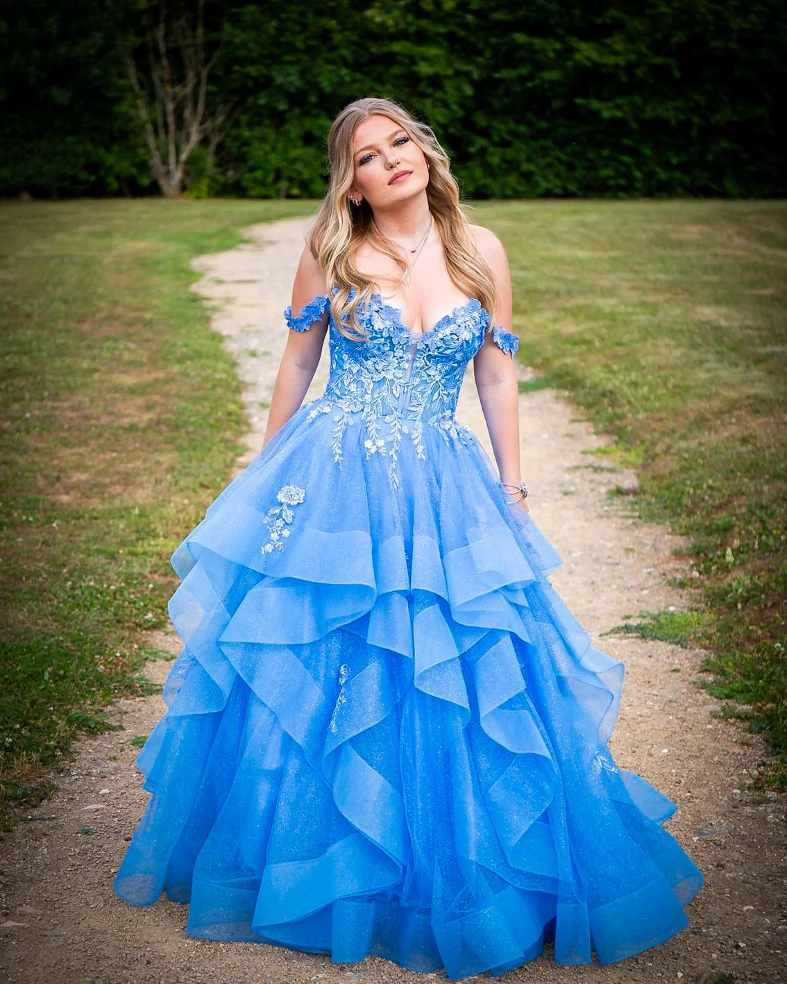 A woman in a bright blue, off-the-shoulder, ruffled ball gown with floral embroidery, standing outdoors on a dirt path with green grass and trees in the background.