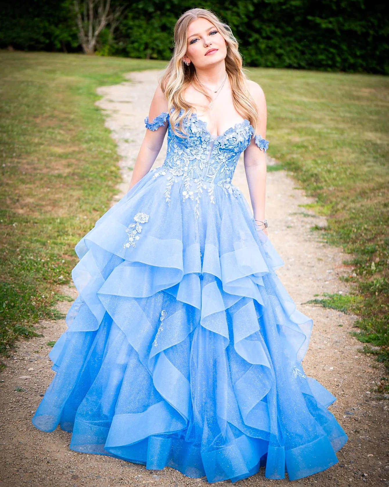 A young woman with blonde hair and light makeup wearing a blue ball gown with lace and floral embroidery, standing on a dirt path in a grassy area with trees in the background.