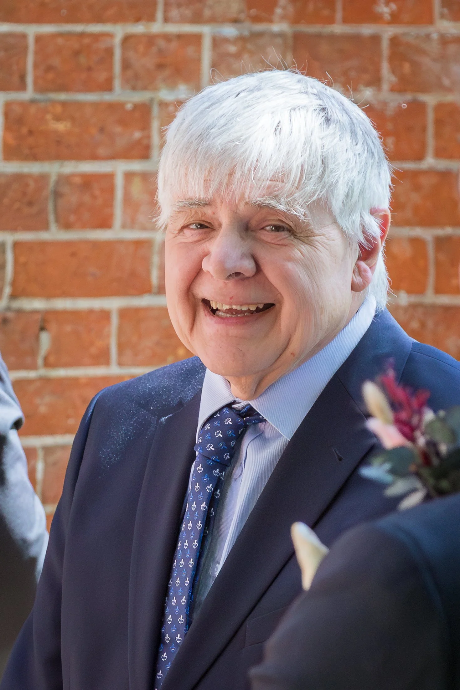 A smiling man with white hair wearing a blue suit, white shirt, and a patterned tie, standing in front of a brick wall.