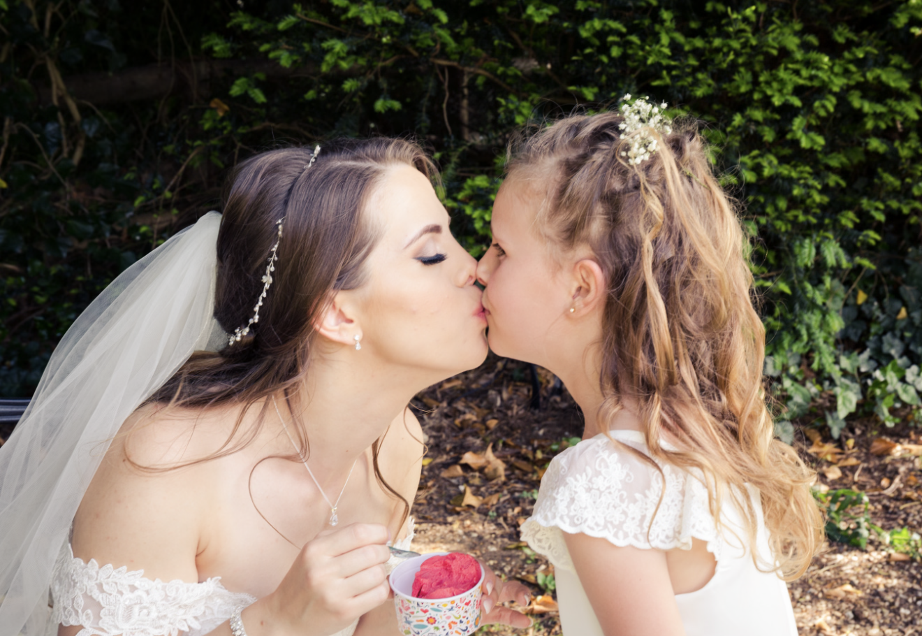 Bride and young girl sharing a kiss outdoors, with greenery in the background. The bride is wearing a white dress with lace details, a veil, and jewelry, and is holding a cup of pink ice cream. The young girl has curly hair decorated with flowers and
