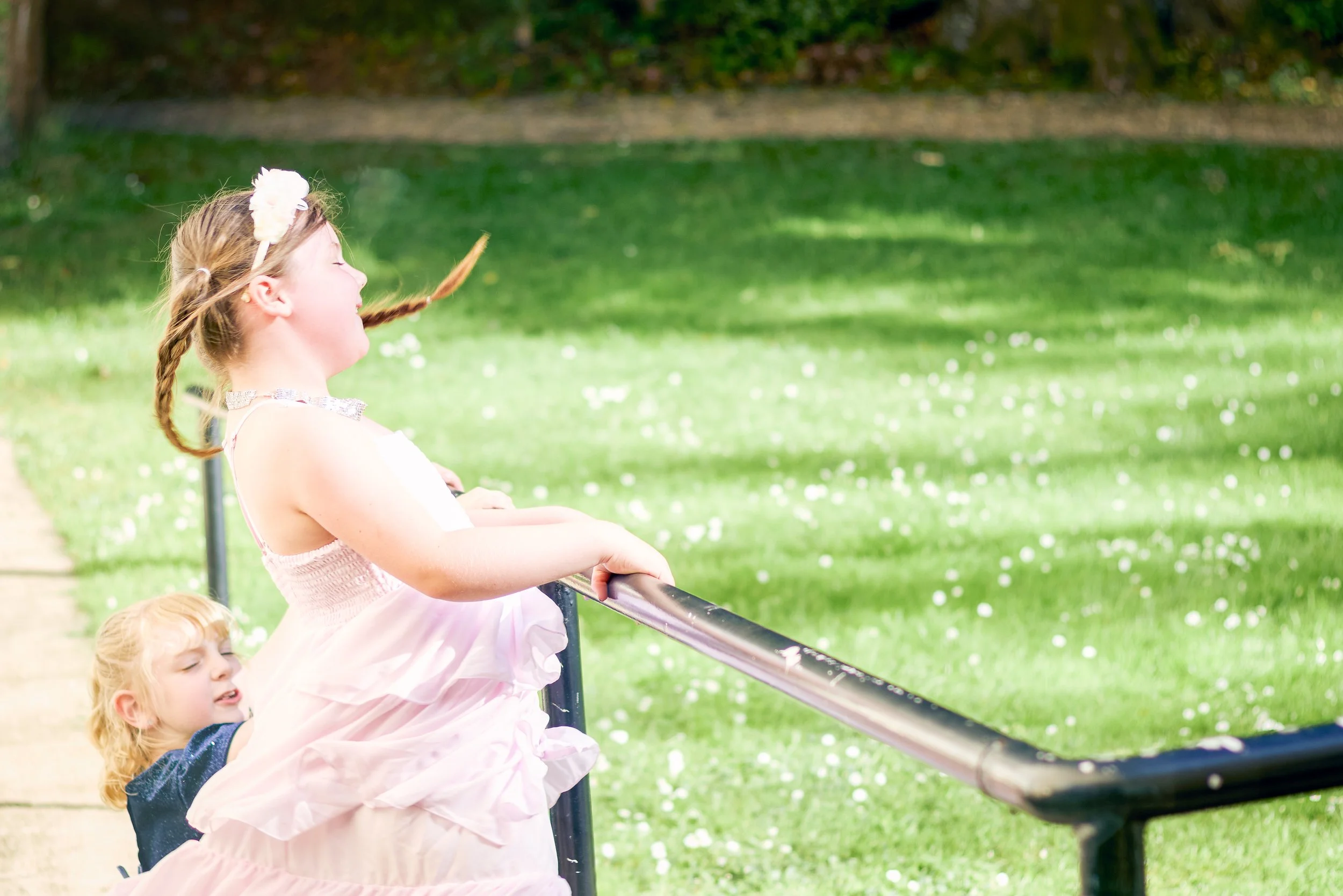 A young girl with blonde hair in braids, wearing a pink dress and a headband with a white flower, is looking over a metal railing in a park, with another girl with curly blonde hair standing behind her. They are surrounded by green grass and trees, w