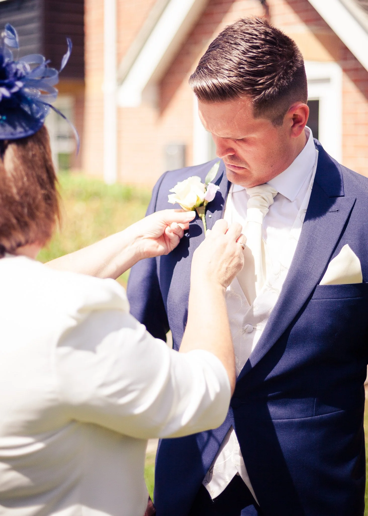 A man in a blue suit and white shirt is being fitted with a white boutonniere by a woman, outdoors in front of a brick building during daytime.