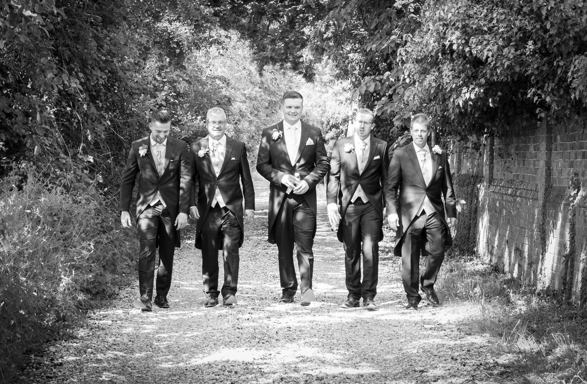 Black and white photo of a group of six men dressed in suits, walking outdoors along a dirt path between trees and a brick wall.