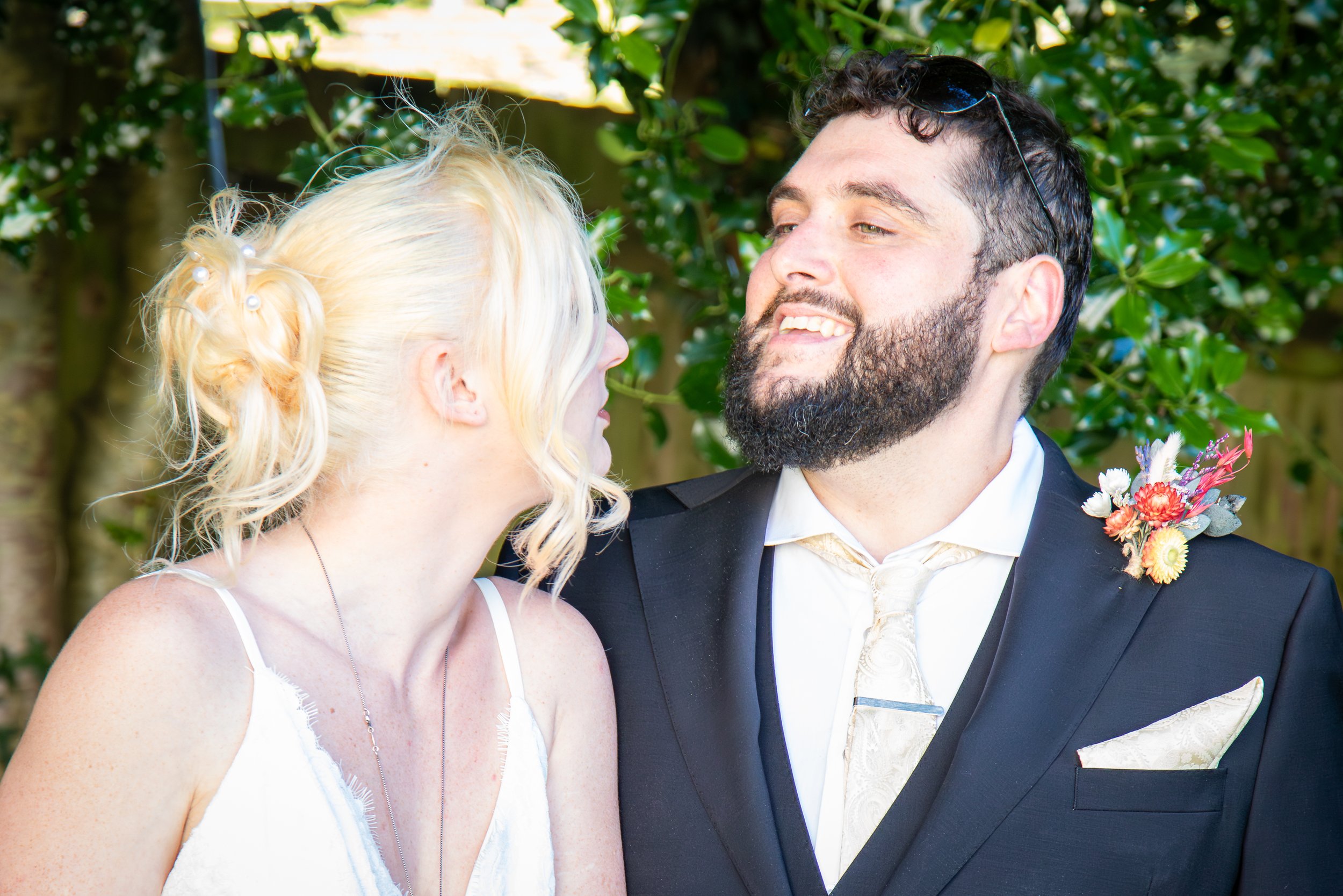 A woman with blonde hair in loose waves and a man with dark hair and a beard standing close together outdoors, dressed in wedding attire with greenery in the background.
