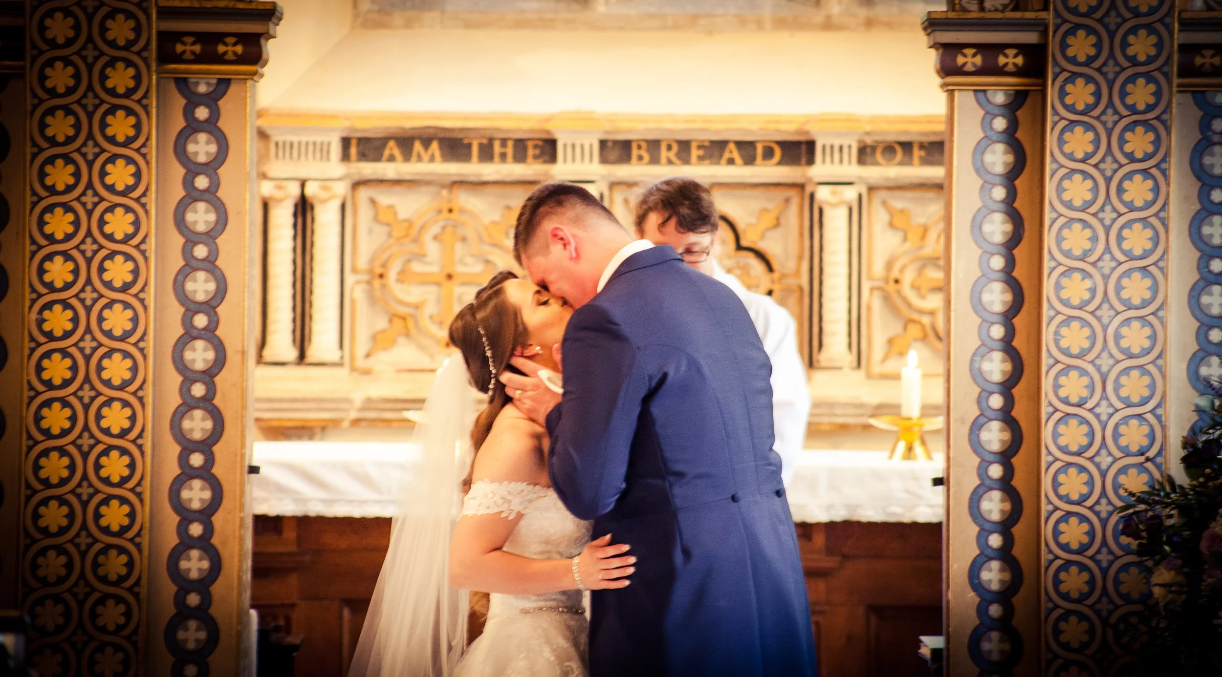 A bride and groom kissing during their wedding ceremony inside a church, with a priest in the background and decorative woodwork framing them.
