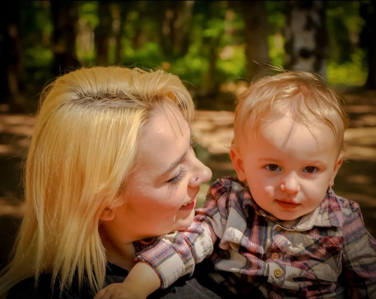 A smiling woman with blonde hair holding a young boy with light hair in an outdoor wooded area.