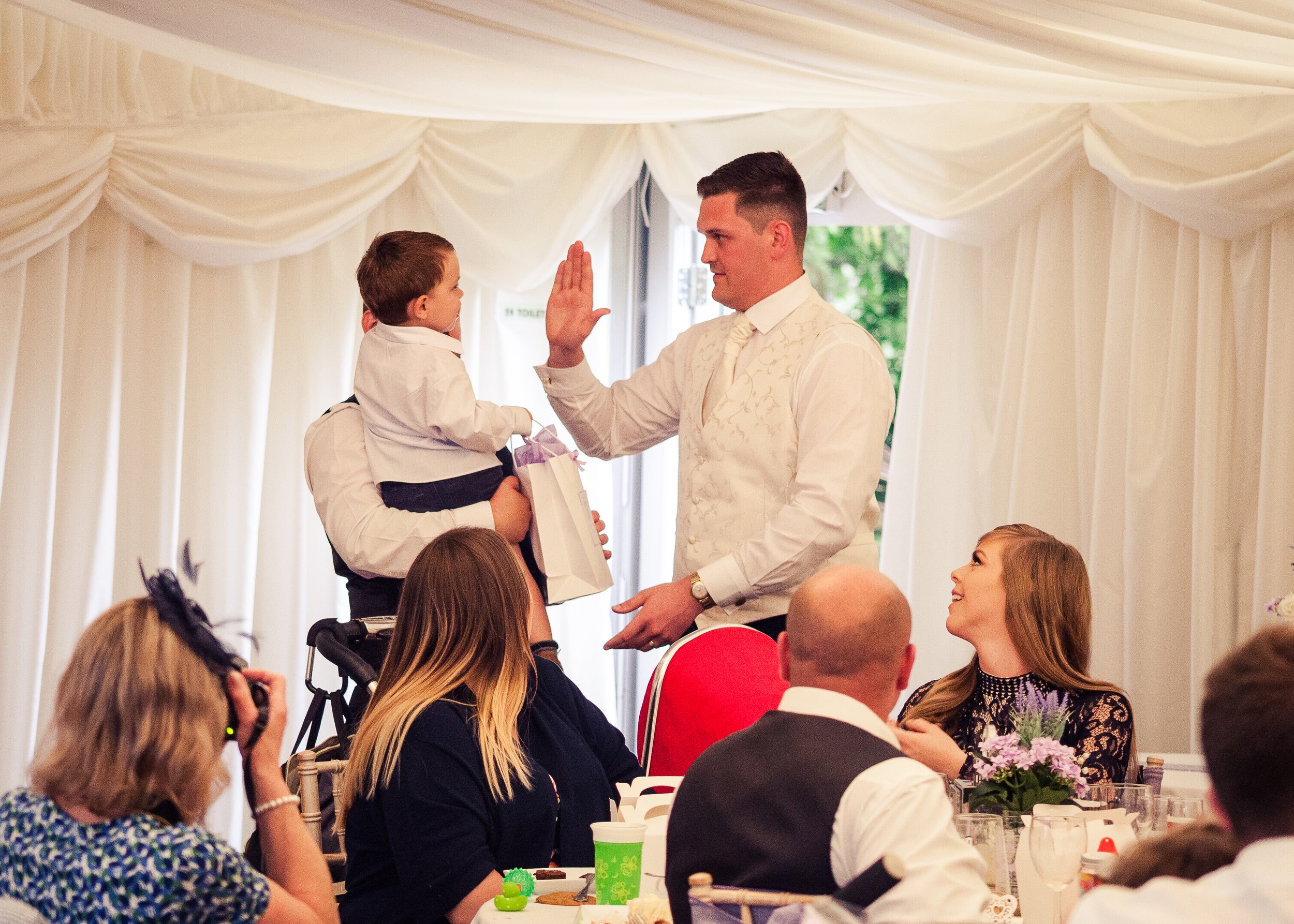 A man in formal attire giving a high five to a young boy at an indoor event with white draped curtains, surrounded by seated attendees.