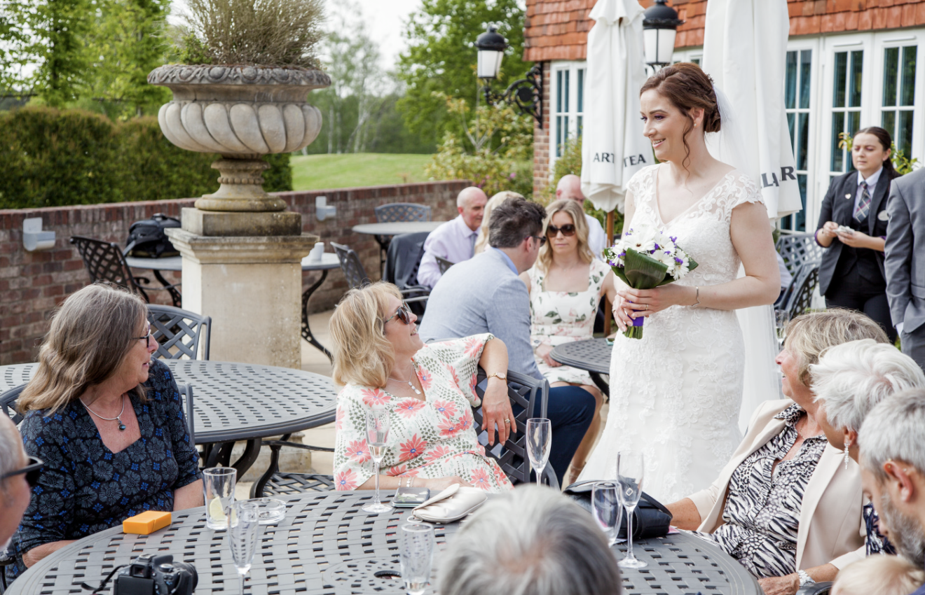 Bride in a white lace wedding dress holding a purple and white bouquet, smiling at guests during a wedding reception outdoors on a patio, with several seated guests and greenery in the background.