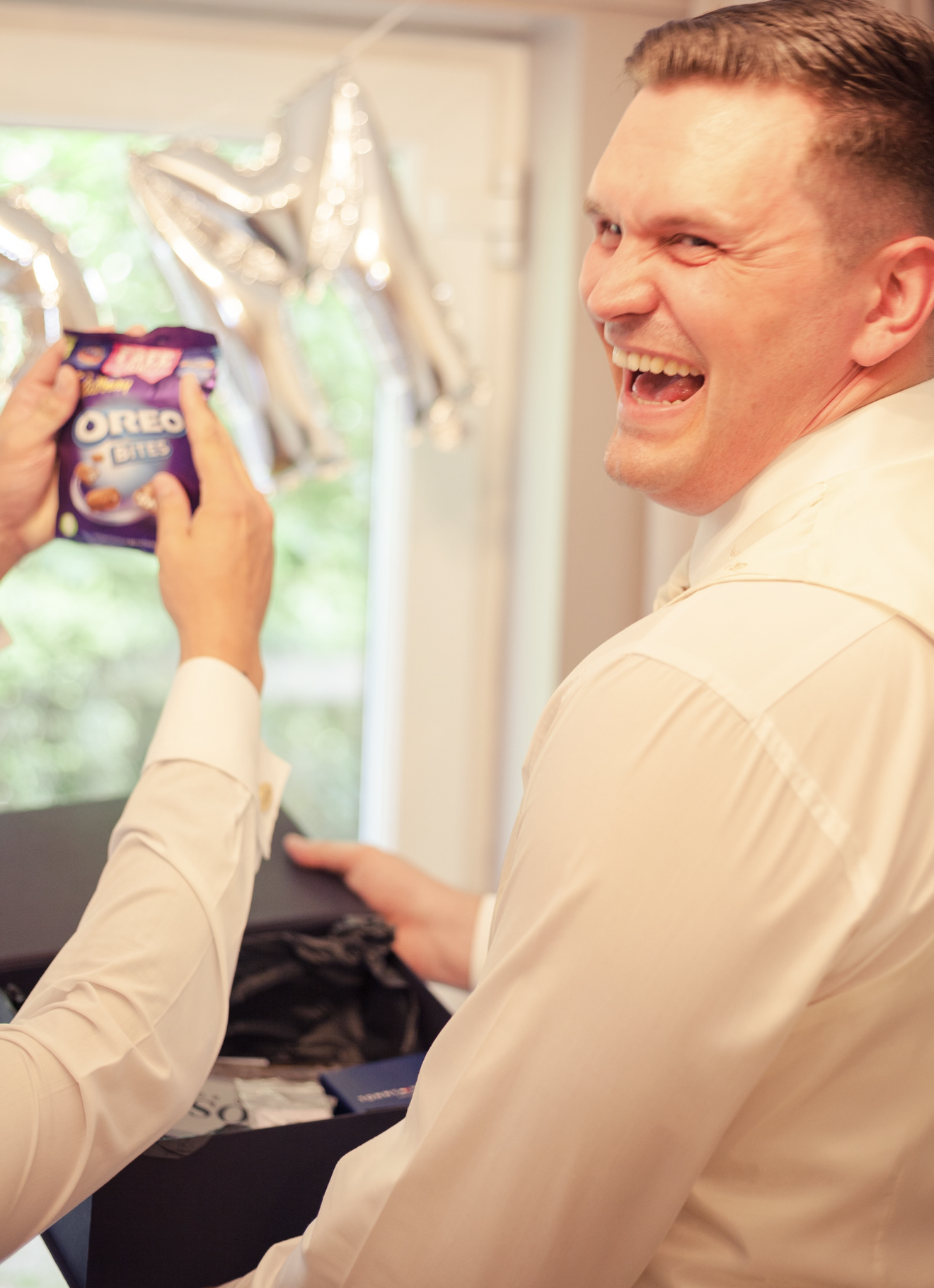 A man in a white shirt laughing while holding a black folder and a person holding a bag of Oreo Bites cookies in front of him, with balloons in the background.