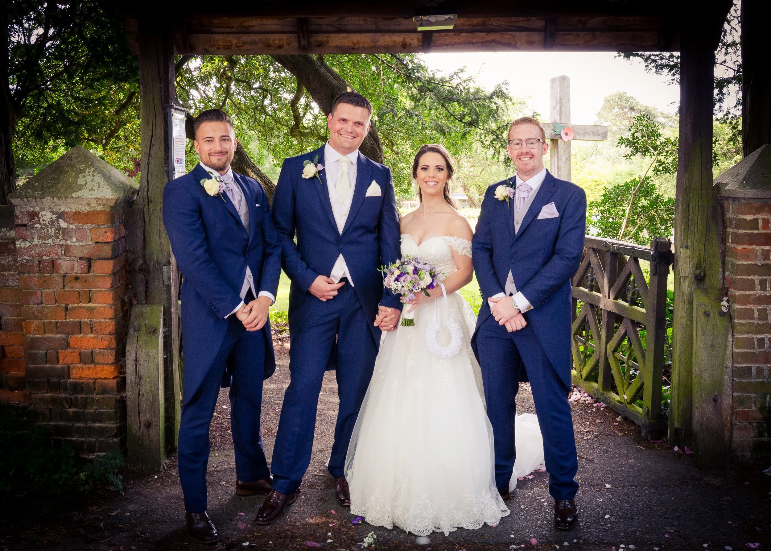 A bride and three groomsmen standing together outdoors under a wooden structure with a cross in the background. The bride is holding a bouquet of flowers, and the groomsmen are dressed in blue suits with floral boutonnieres. The scene appears to be d