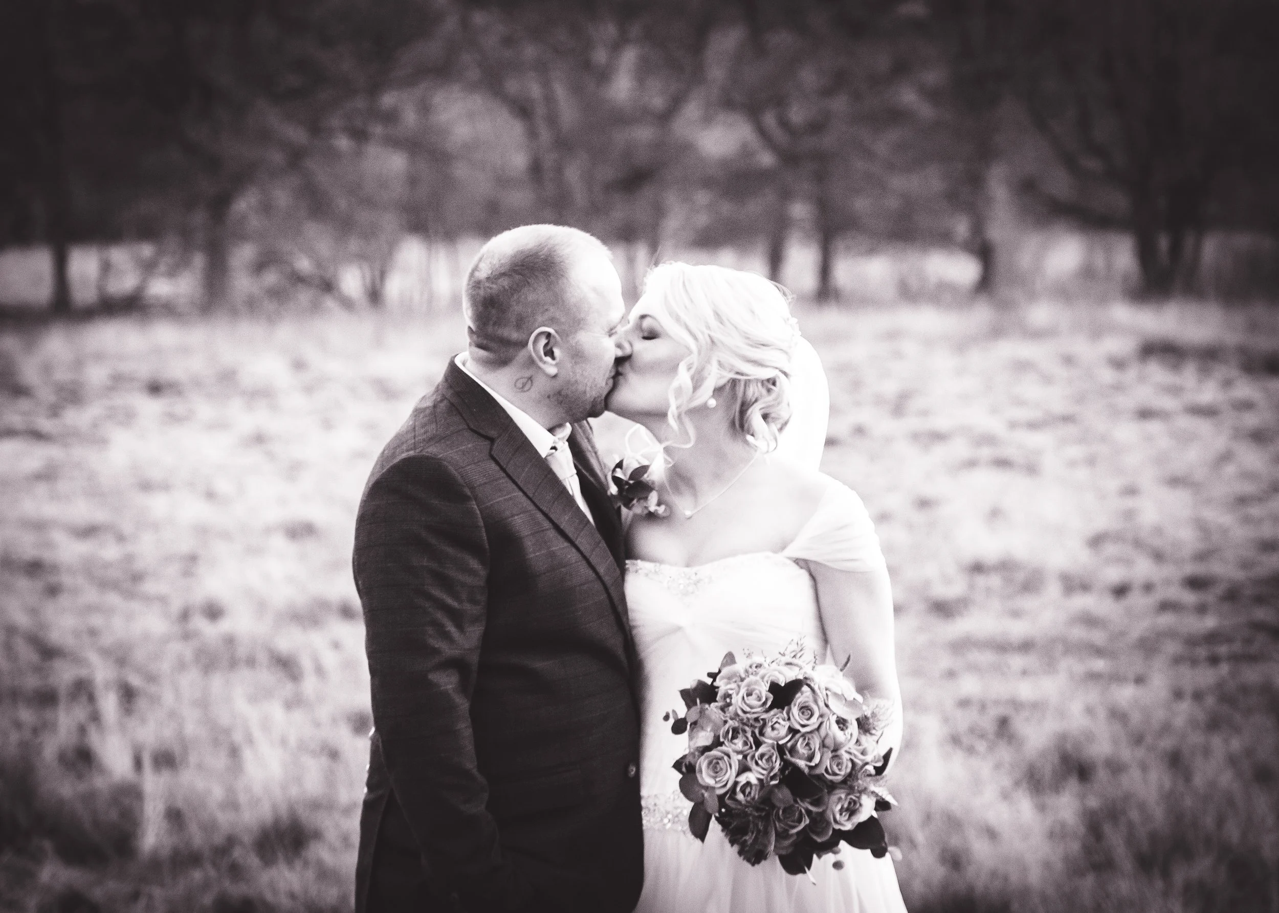 A black and white photo of a bride and groom kissing outdoors during their wedding, with trees in the background.
