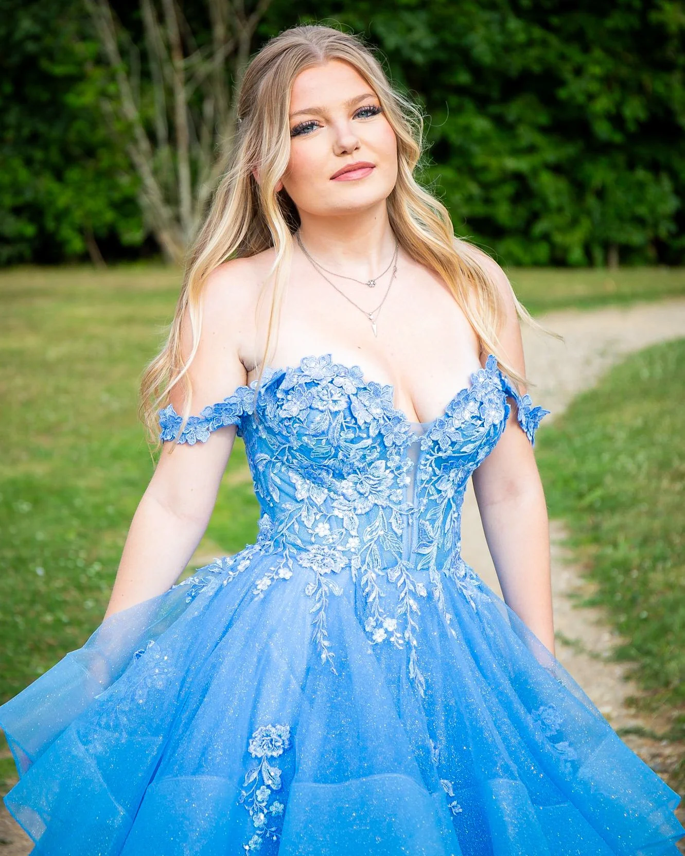 A young woman with long blonde hair standing outdoors in a green park, wearing a blue floral off-shoulder princess gown with lace and floral embroidery, accessorized with layered necklaces.